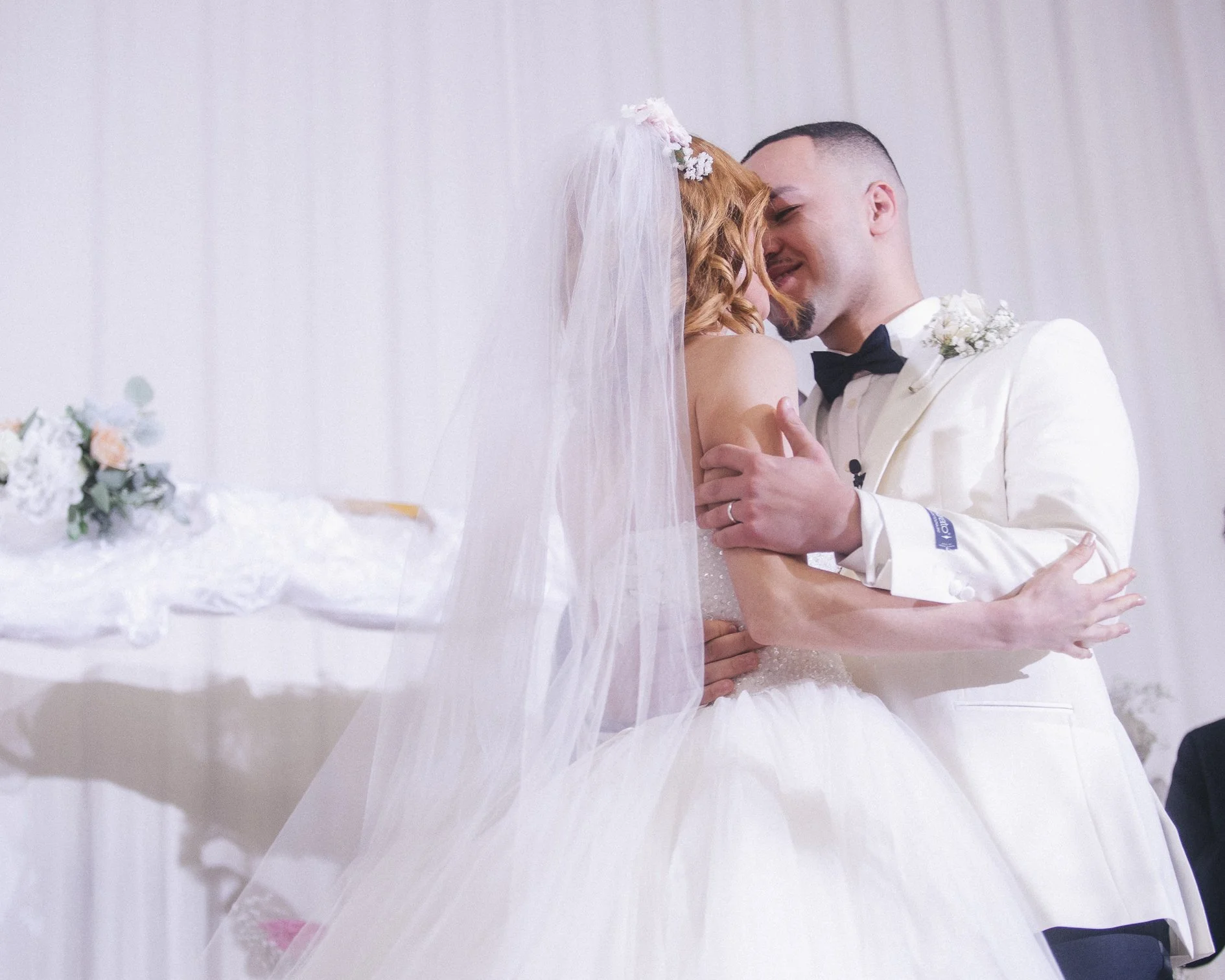A bride and groom share a dance during their wedding, with the bride in a white wedding gown and veil, and the groom in a white tuxedo with a black bow tie, indoors with white drapes.