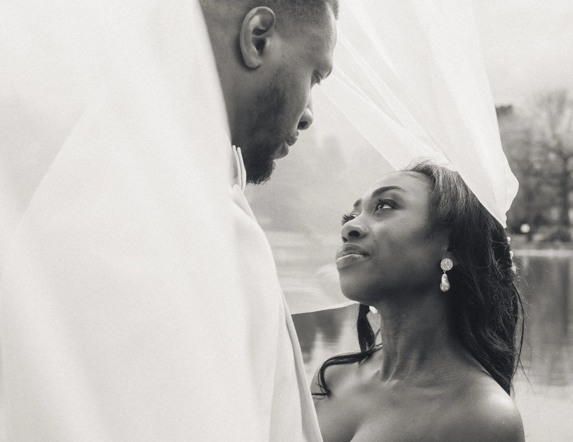 Black and white photo of a couple standing close together outdoors, with the woman looking up at the man who is partially wrapped in the brides vail, both gazing into each other's eyes.