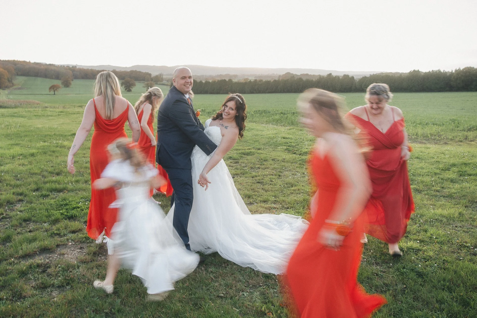 A wedding party dancing outdoors on a grassy field, with a bride and groom in the center surrounded by bridesmaids and a flower girl, all dressed in vibrant red and white dresses, against a rural landscape with rolling hills.