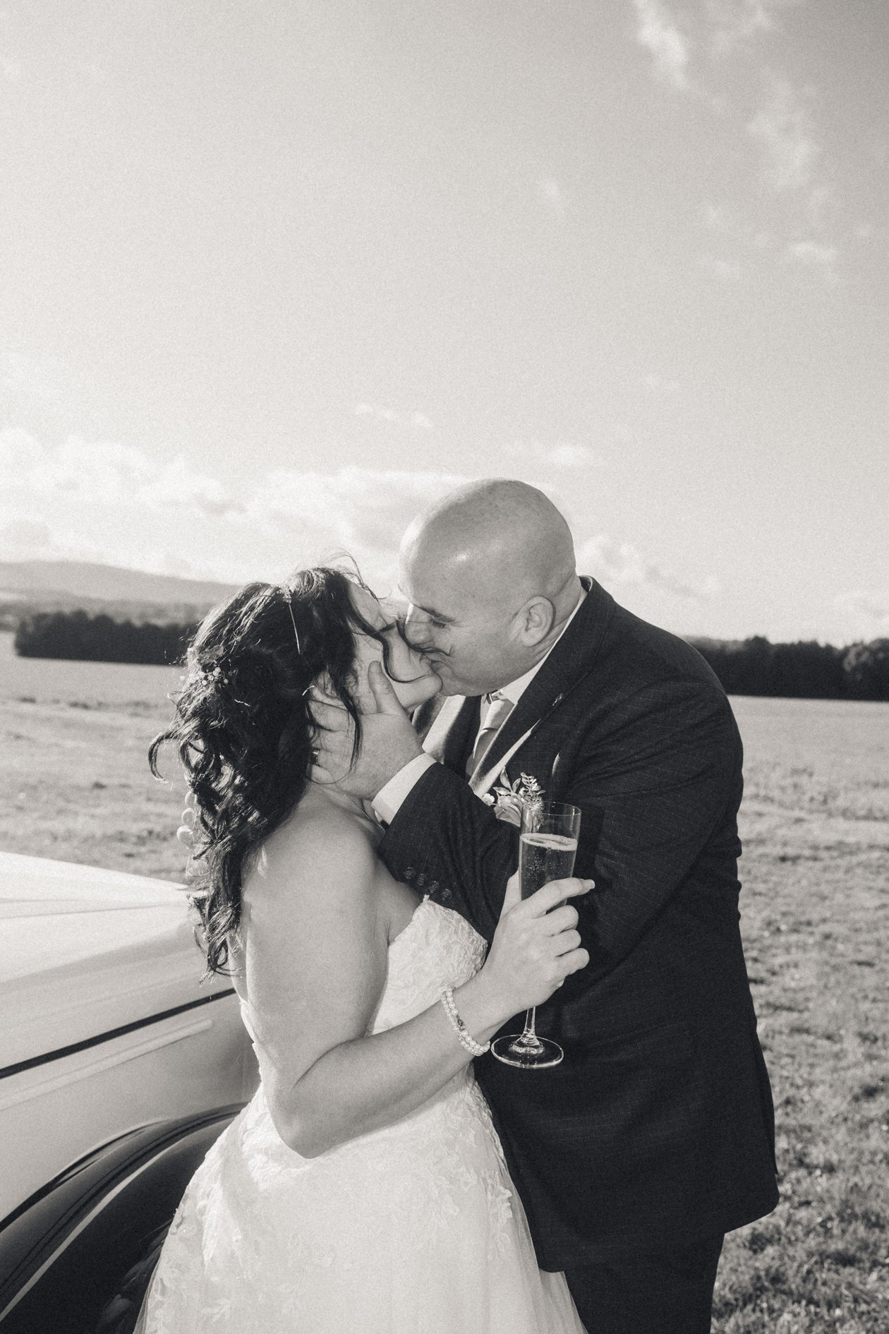 A black and white photo of a newlywed couple kissing outdoors with a car in the background. The bride is holding a champagne flute and wearing a wedding dress and bracelet, while the groom is in a suit.