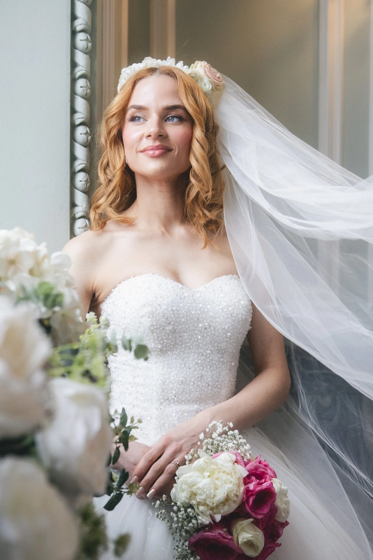 A bride with red hair in a white wedding dress and veil standing next to a window, holding a bouquet of pink and white flowers.