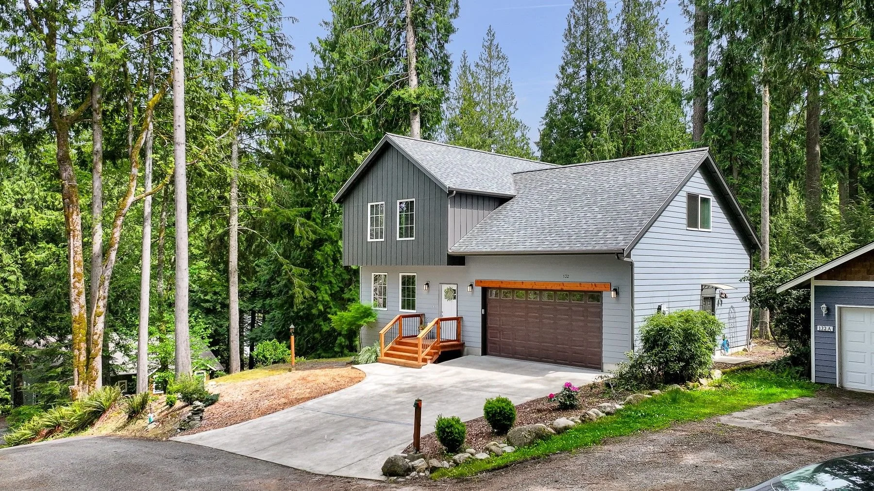 A two-story gray house with a brown garage door, small front porch with wooden steps, and surrounded by tall trees and greenery in Billingham WA