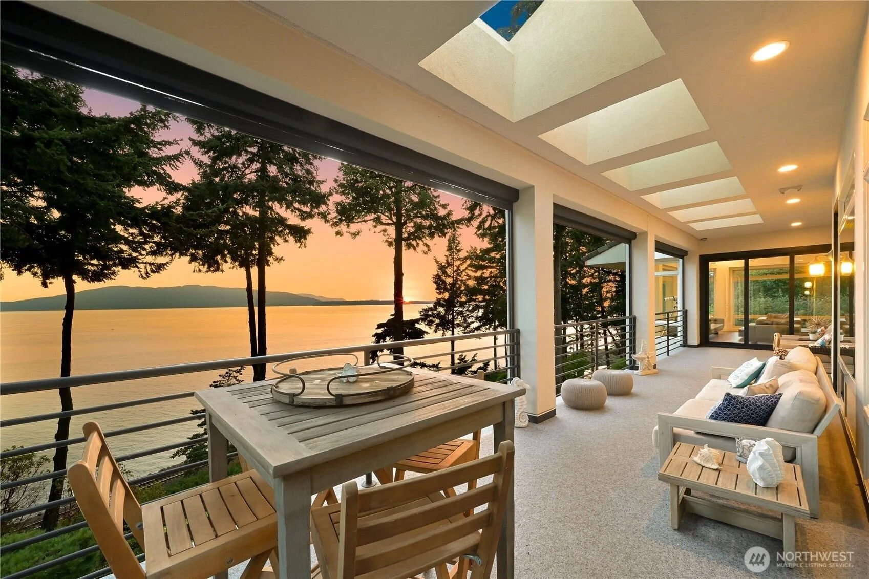 Covered balcony with outdoor seating, overlooking a body of water with trees and mountains at sunset. In Bellingham WA