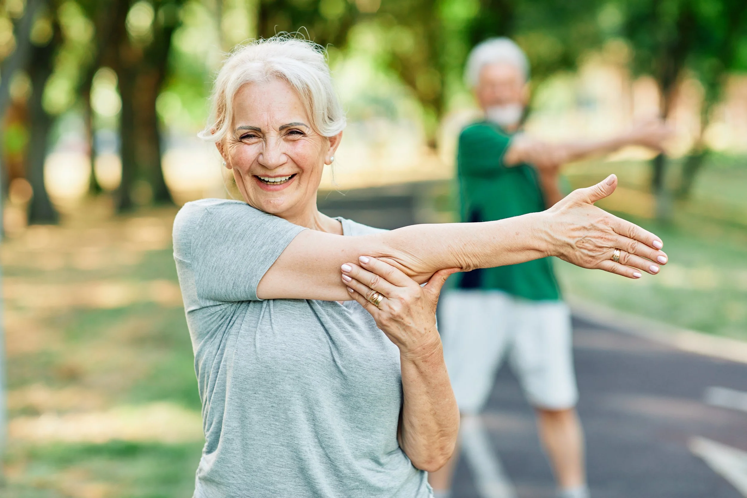Smiling elderly woman in gray shirt stretching arm outdoors with a happy expression, elderly man in green shirt and white shorts doing similar stretch in the background at the park.
