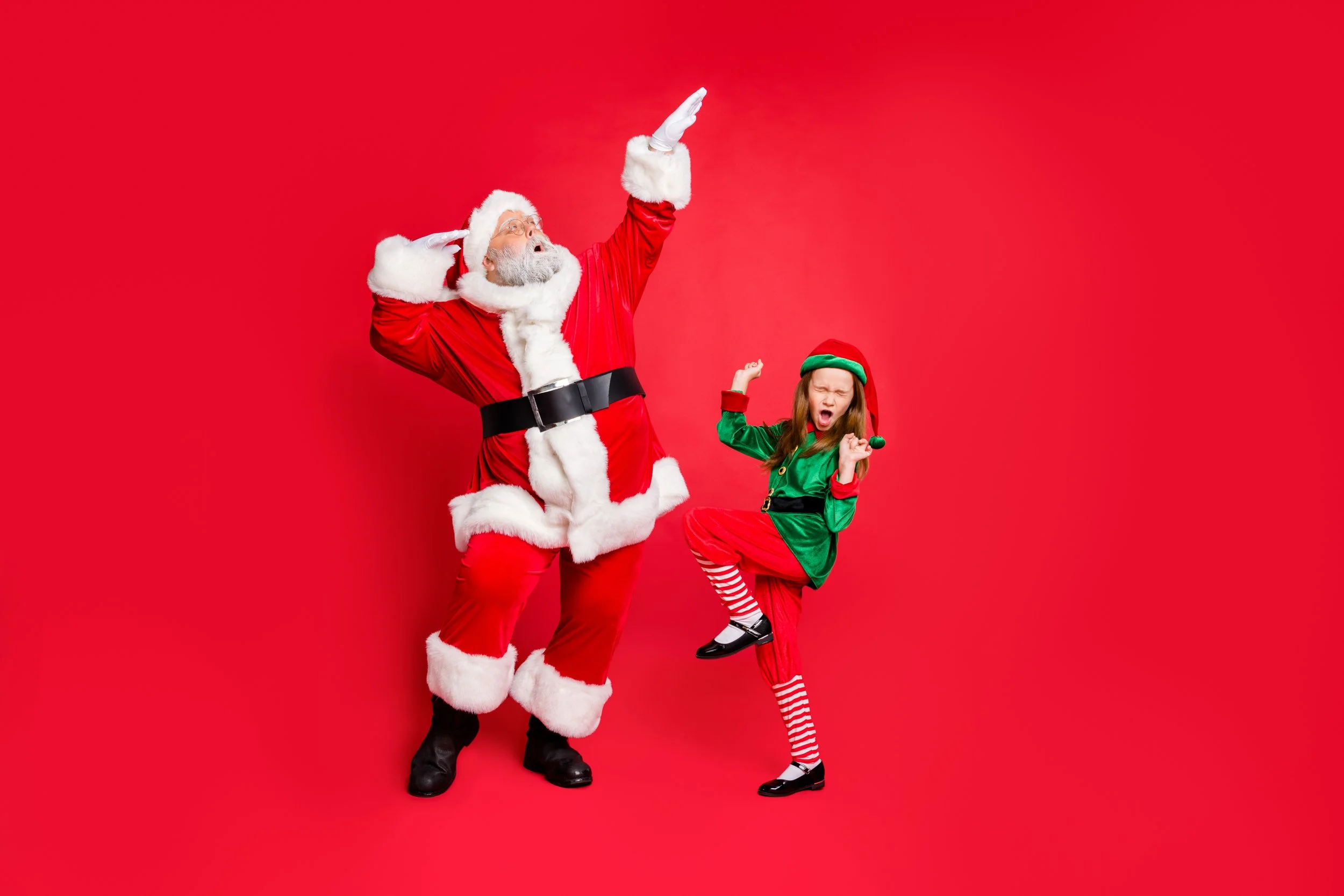 Santa Claus with a white beard and red suit poses with a young girl dressed as an elf, both making playful gestures against a red background.