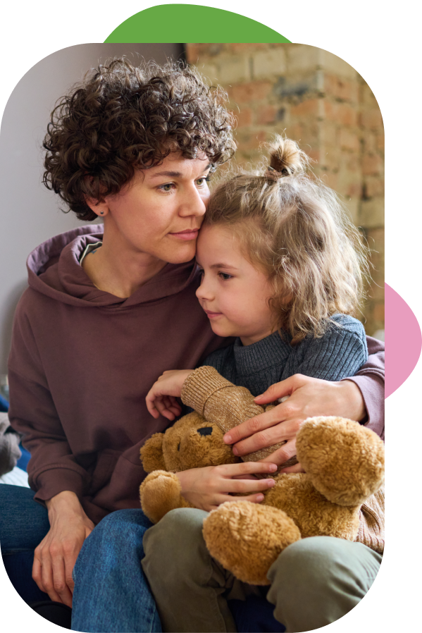 A woman and a young girl sitting close together, cuddling a teddy bear in a cozy setting with exposed brick wall in the background.