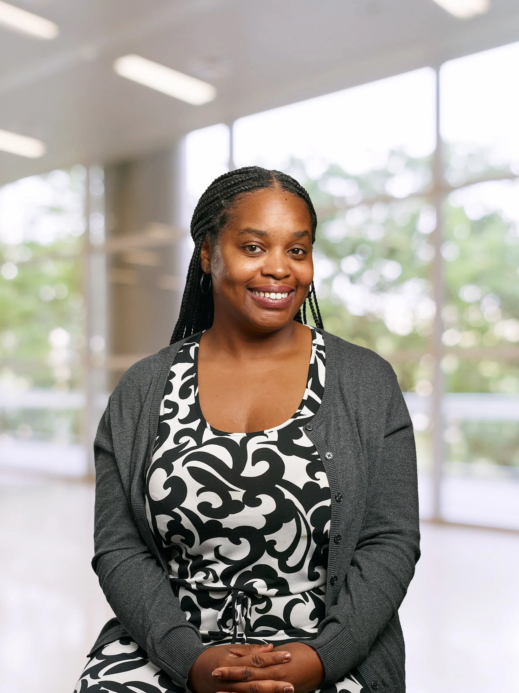 Portrait of a smiling woman with braided hair, wearing a patterned dress and gray cardigan, sitting indoors with large windows and greenery in the background.