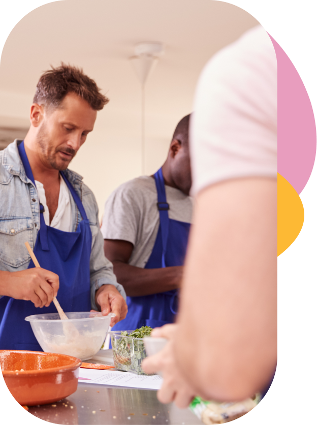 People cooking together in a kitchen, with a man mixing ingredients in a bowl and others preparing food nearby.