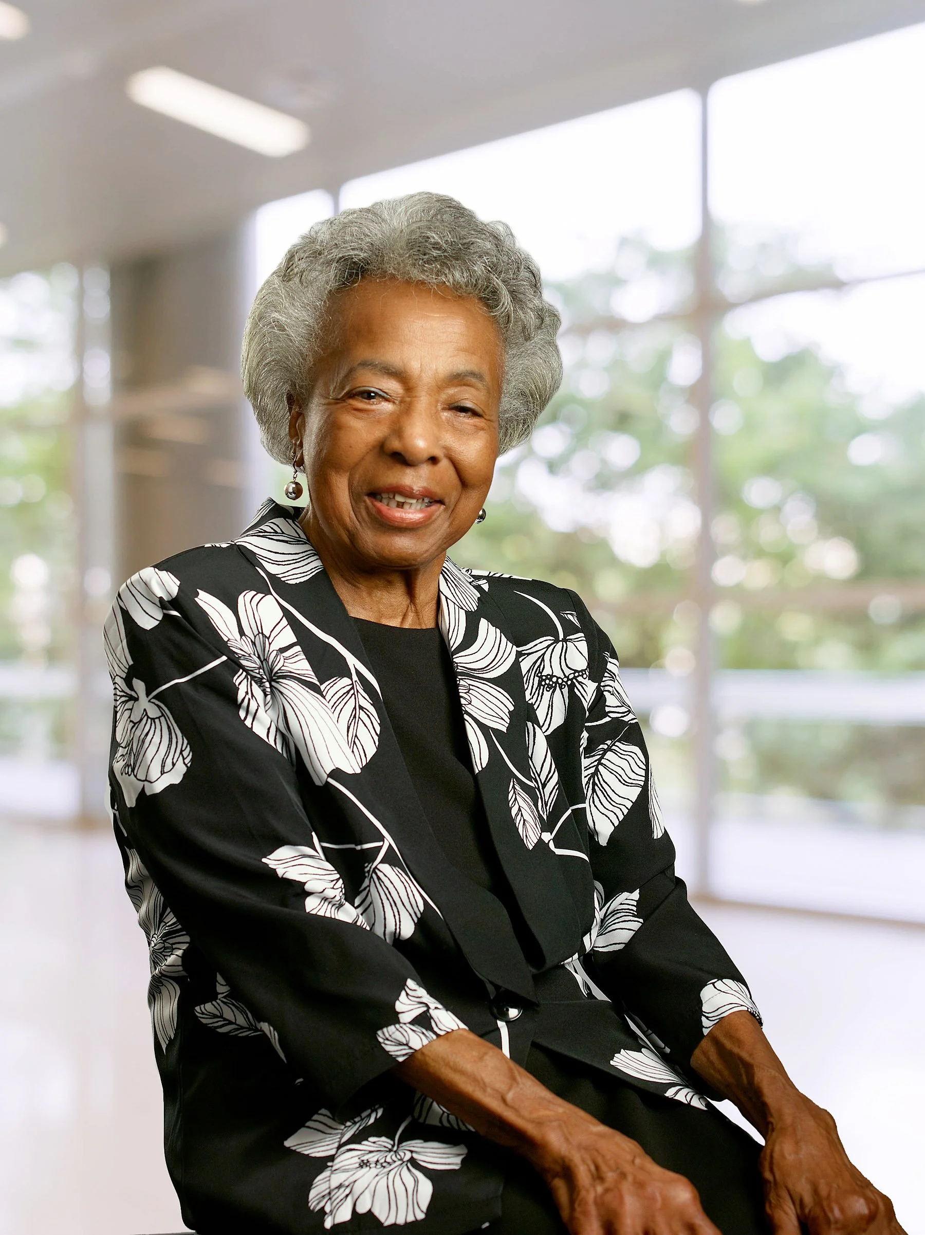 Portrait of an elderly African American woman with gray hair, smiling, wearing a black and white floral blazer, sitting in a modern indoor setting with large windows in the background.