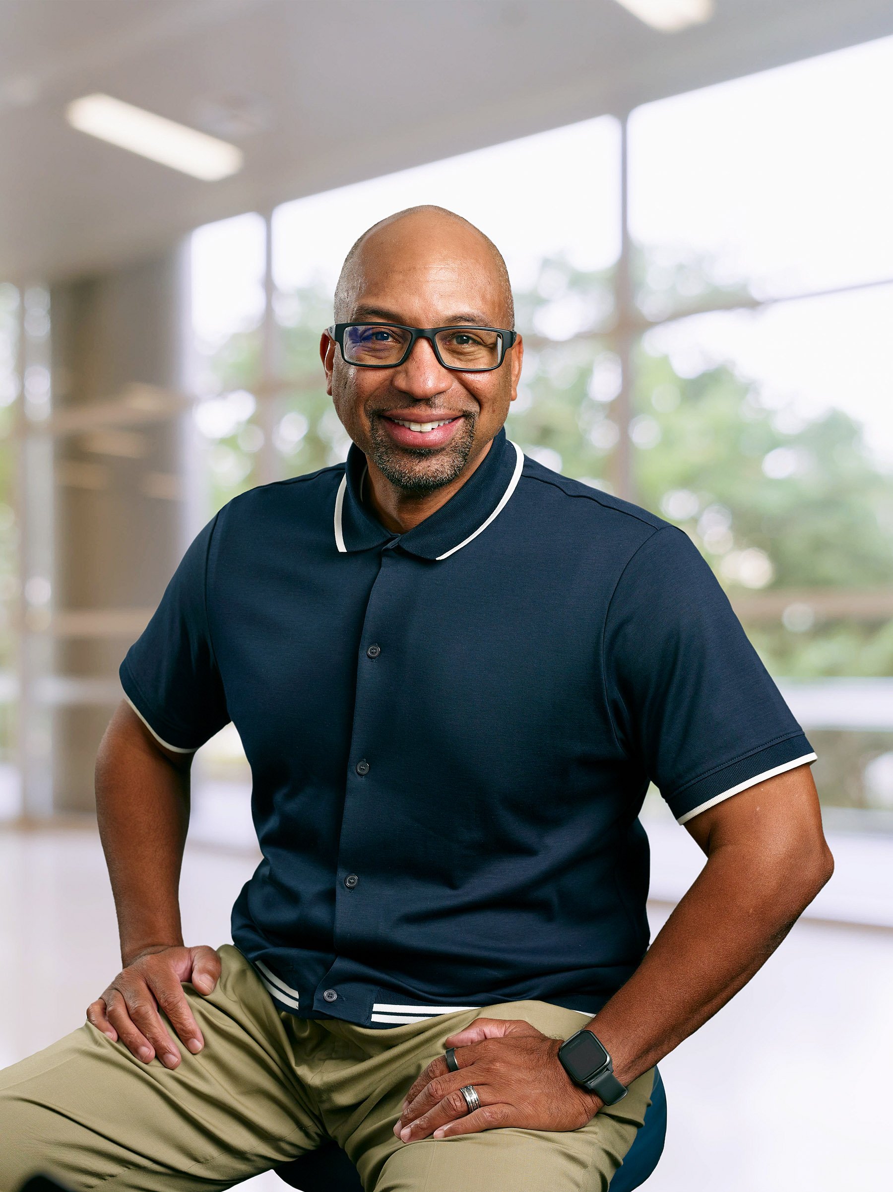 Smiling middle-aged man with glasses, bald, wearing a navy polo shirt and khaki pants, seated indoors with large windows showing trees outside.