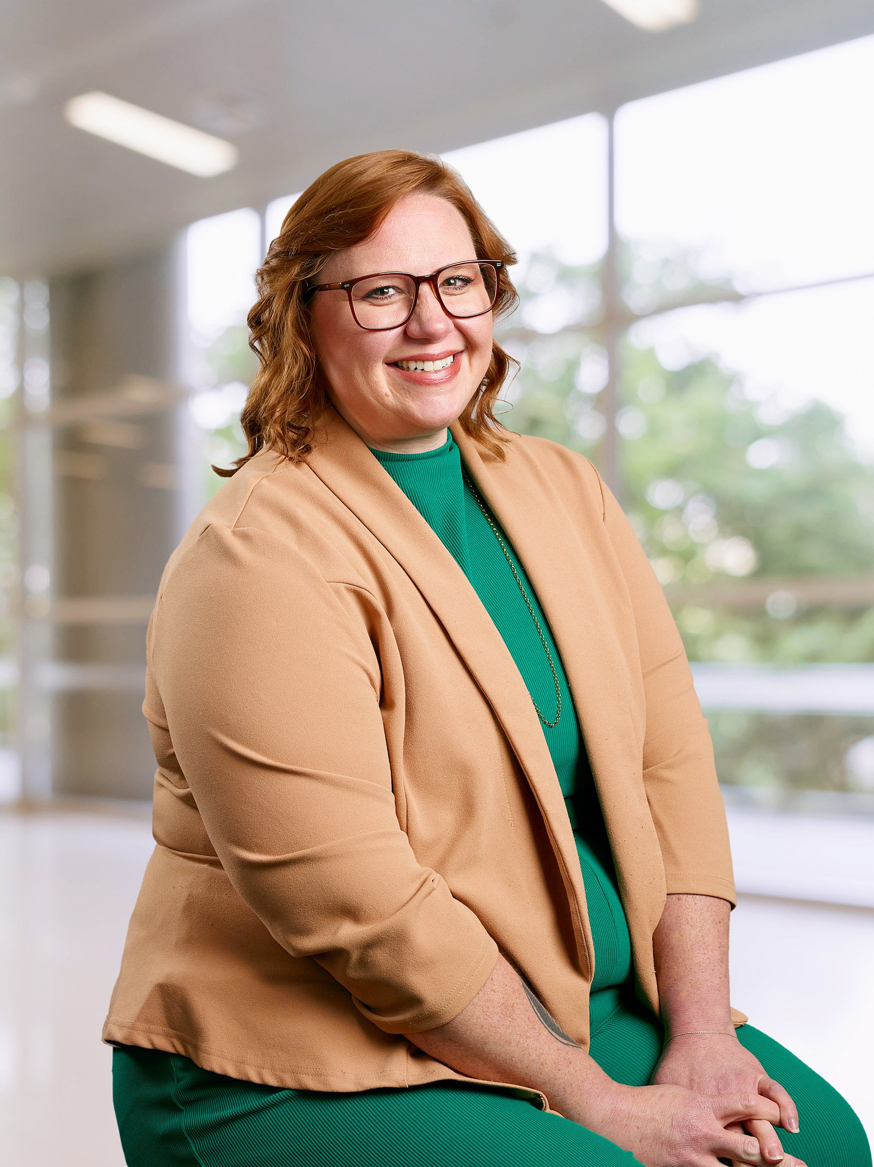 A woman with red hair, glasses, and a warm smile, dressed in a tan blazer and green top, sitting indoors with large windows showing greenery outside.