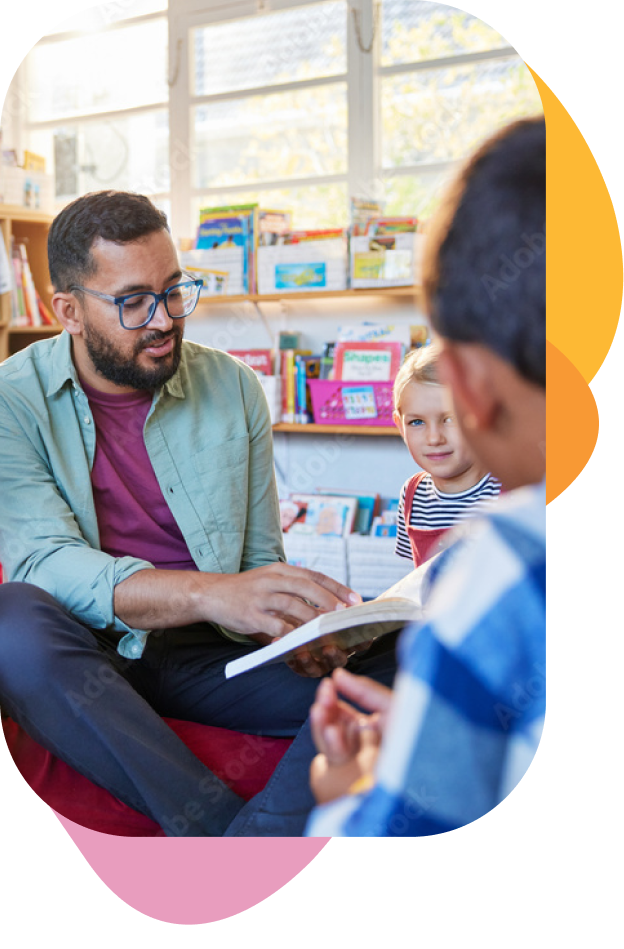 A man in glasses and a green shirt reading a book to two children in a colorful classroom