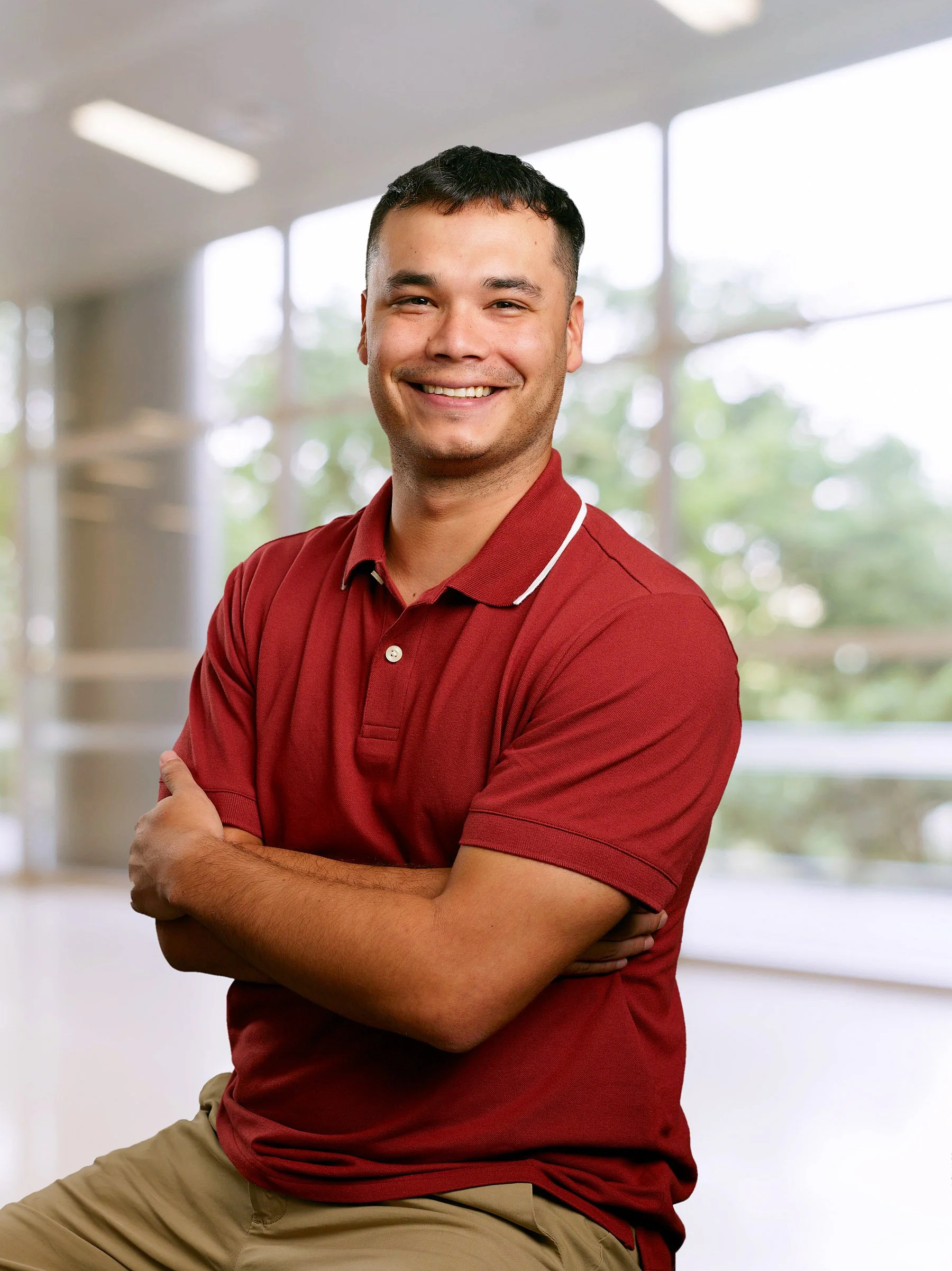 Smiling man in red polo shirt with crossed arms, sitting indoors with large windows and greenery outside.