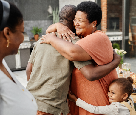 Three people hugging warmly in a cozy kitchen, with a woman smiling happily while a young girl watches.