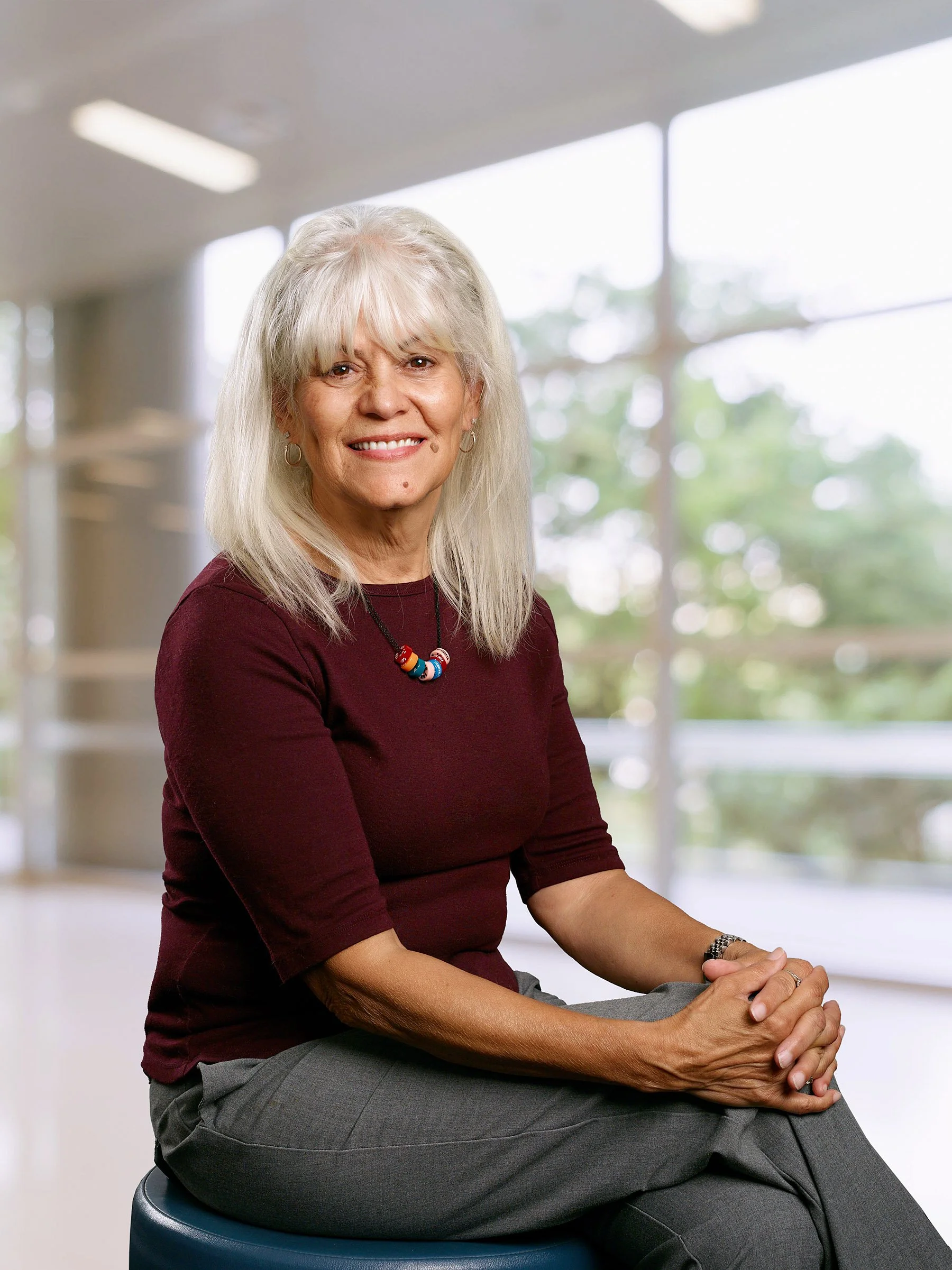 A smiling elderly woman with gray hair and light skin, wearing a maroon top, gray pants, a colorful beaded necklace, and a watch, sitting indoors on a blue stool near large windows with a view of green trees outside.