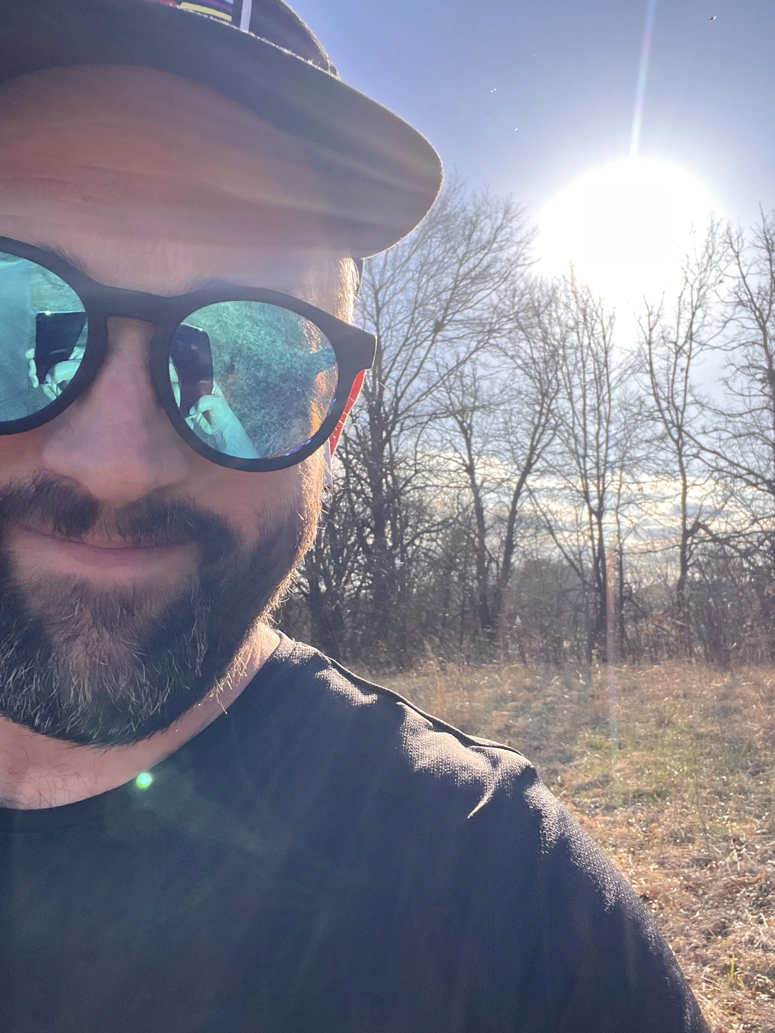 A person with sunglasses and a cap taking a selfie outdoors on a sunny day with bare trees in the background.