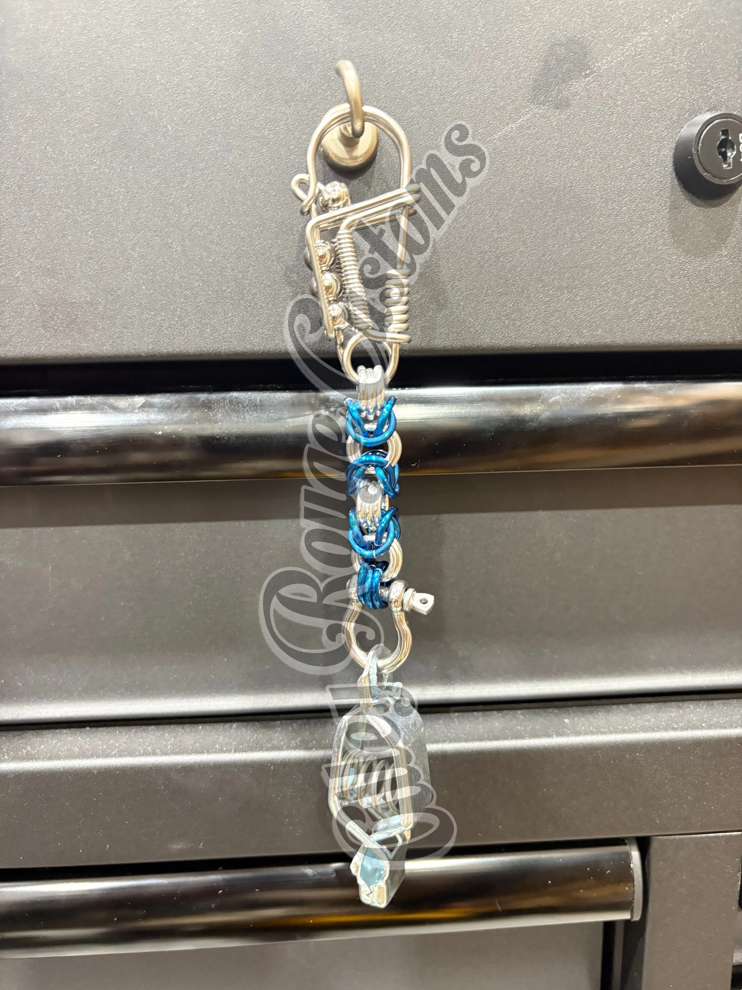 A silver safety pin holding a keychain with blue and silver links and a silver clamp, hanging on a gray metal surface.