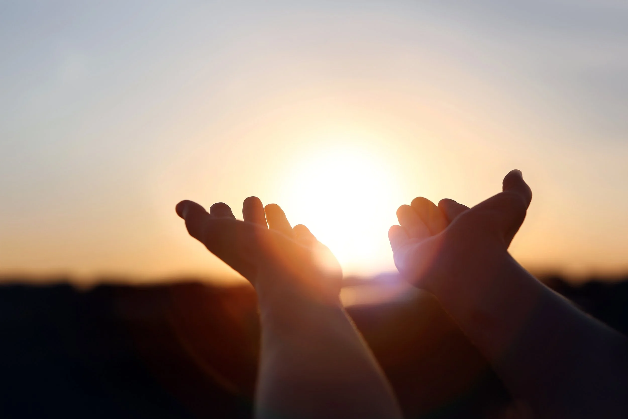 Two hands reaching towards the setting sun during sunset or sunrise.