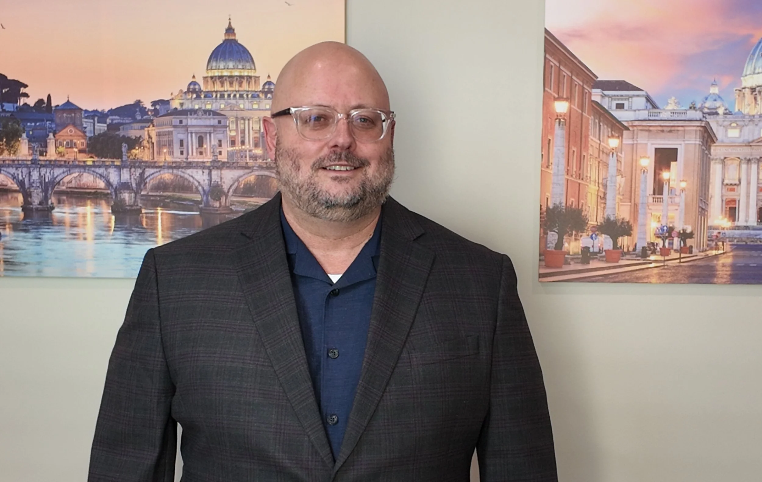 Mario Uribe, therapist, poses with glasses and beard in front of artworks depicting European cityscapes with domed buildings and bridges during sunset.