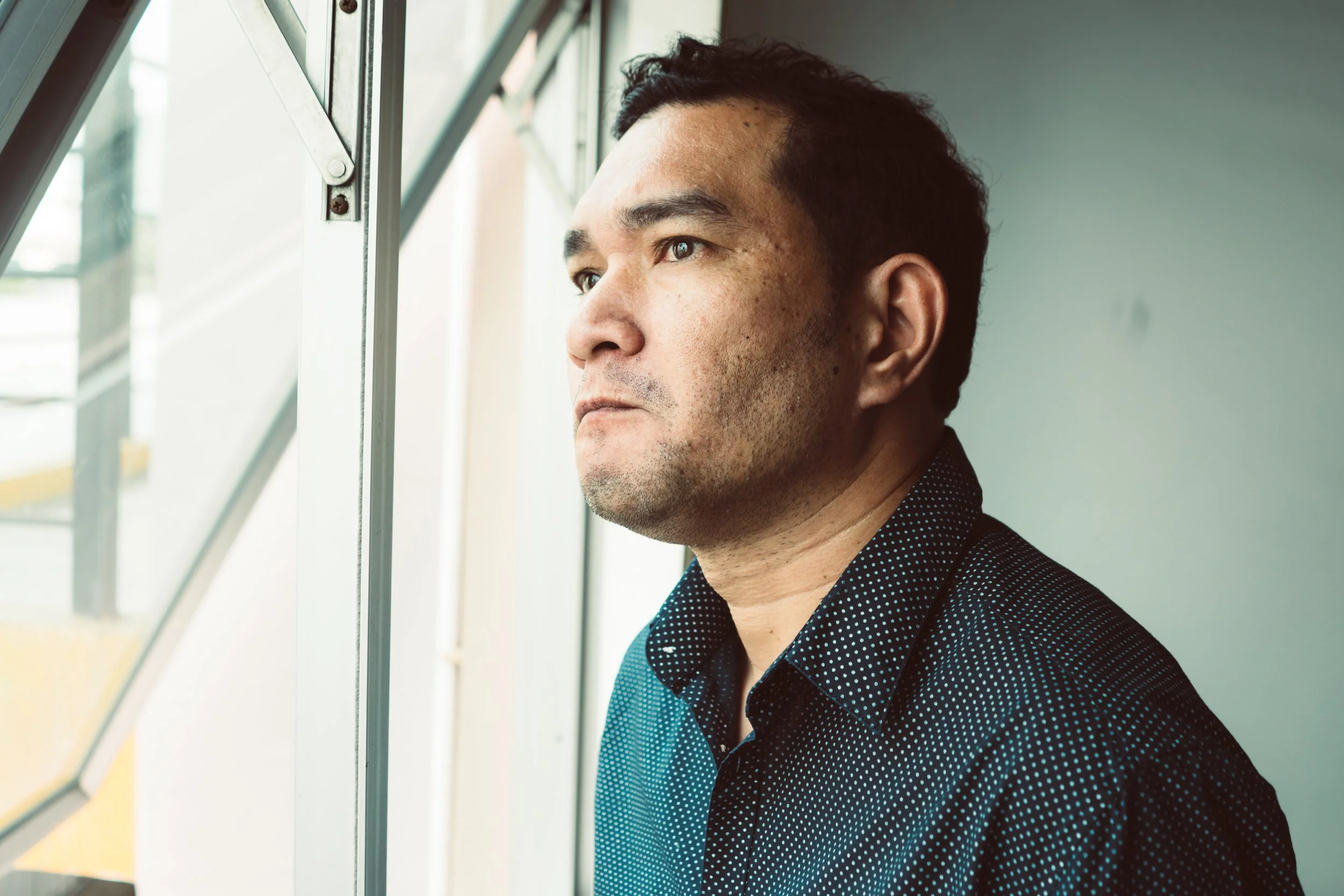 A man in a dark dotted shirt looking out a window, with a serious expression.
