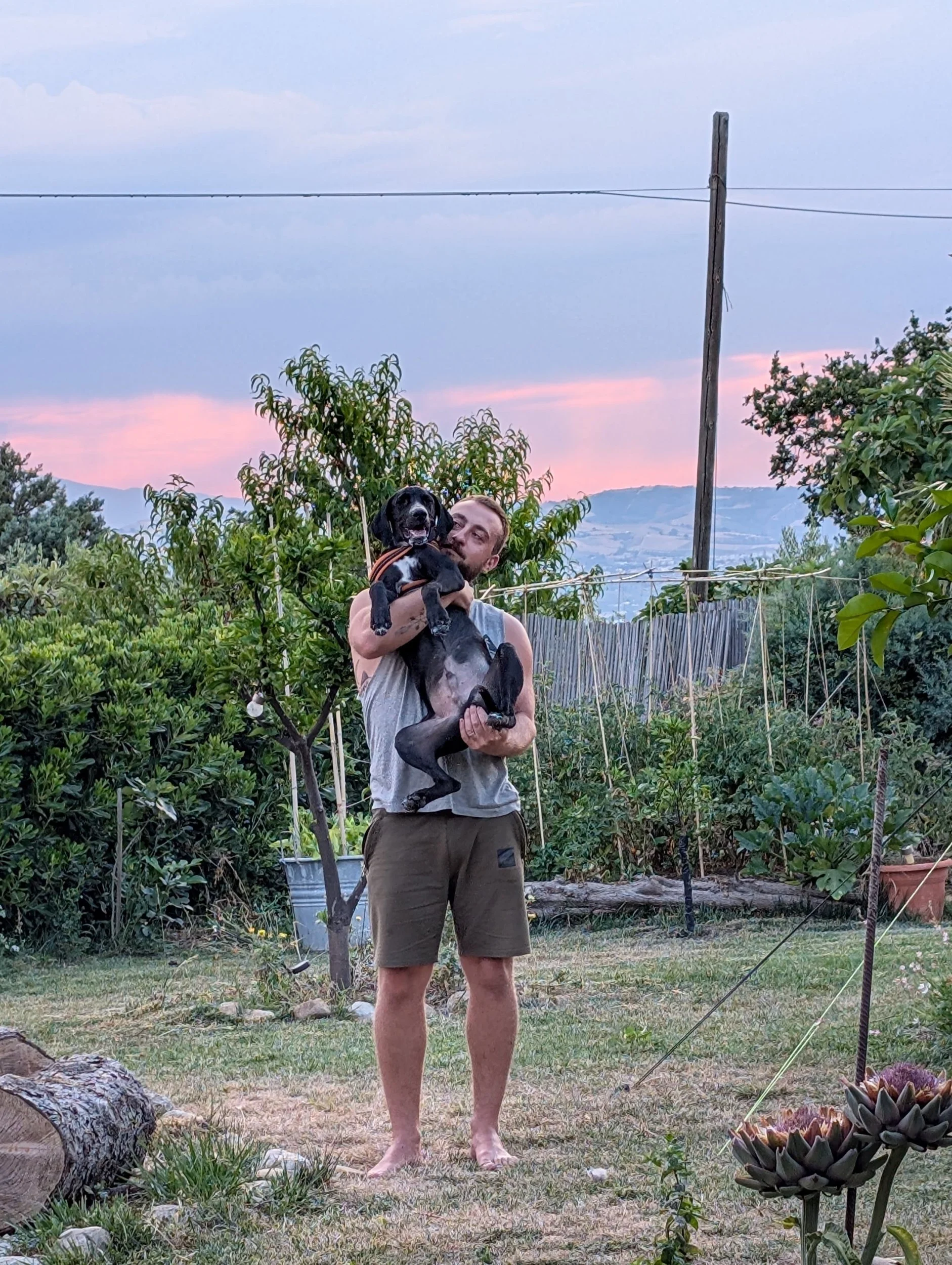 Ein Mann hält einen schwarzen Hund in einem Garten bei Sonnenuntergang. Im Hintergrund sind Bäume, Blumen und eine Hütte sichtbar.