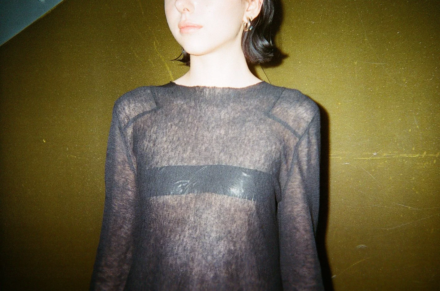 A woman wearing a sheer black top with a black bandeau underneath, standing against a dark yellow background.