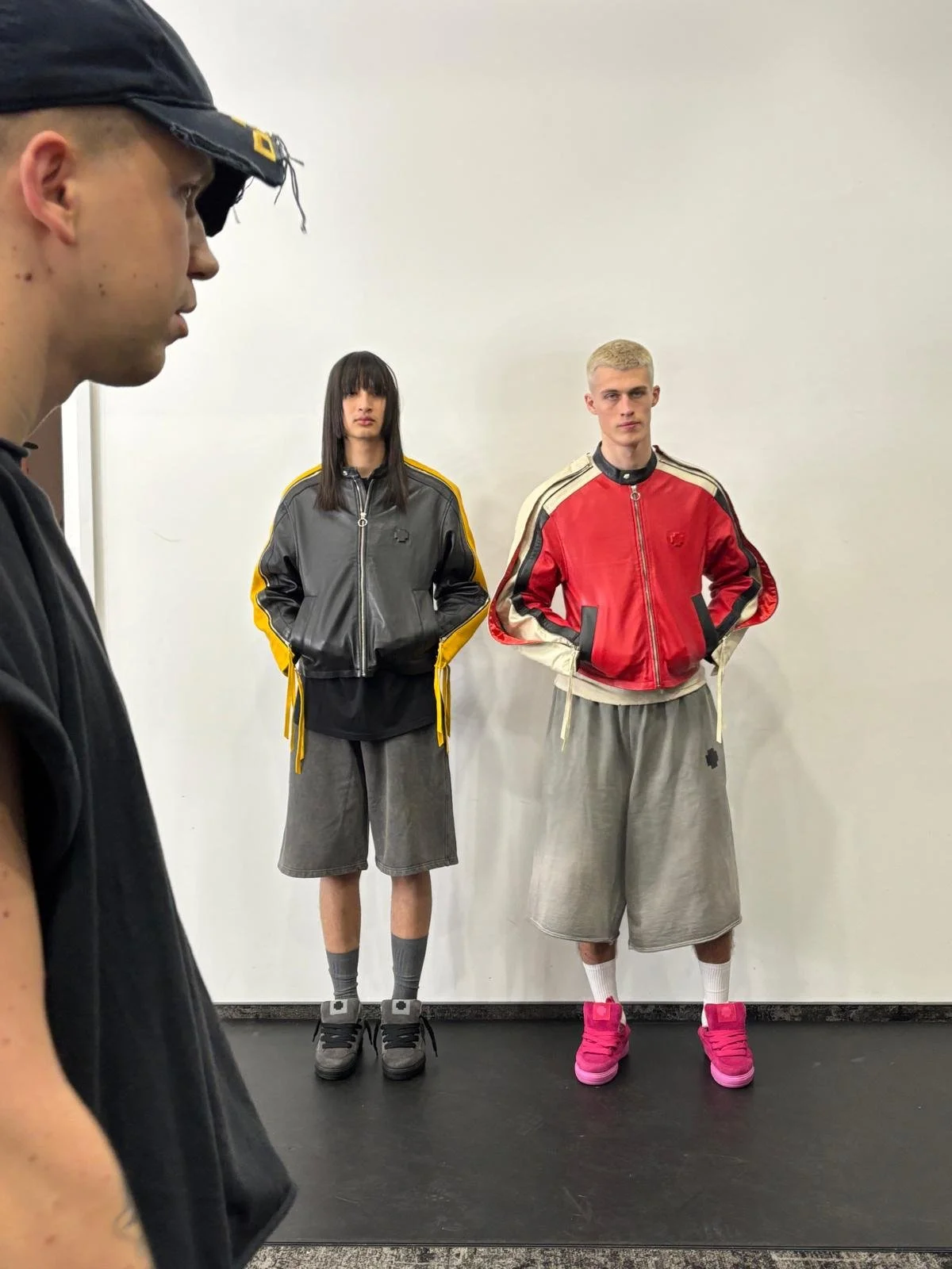 Two young men and one young woman standing against a plain white wall, all wearing sporty track jackets and shorts, with the man on the right wearing bright pink shoes.