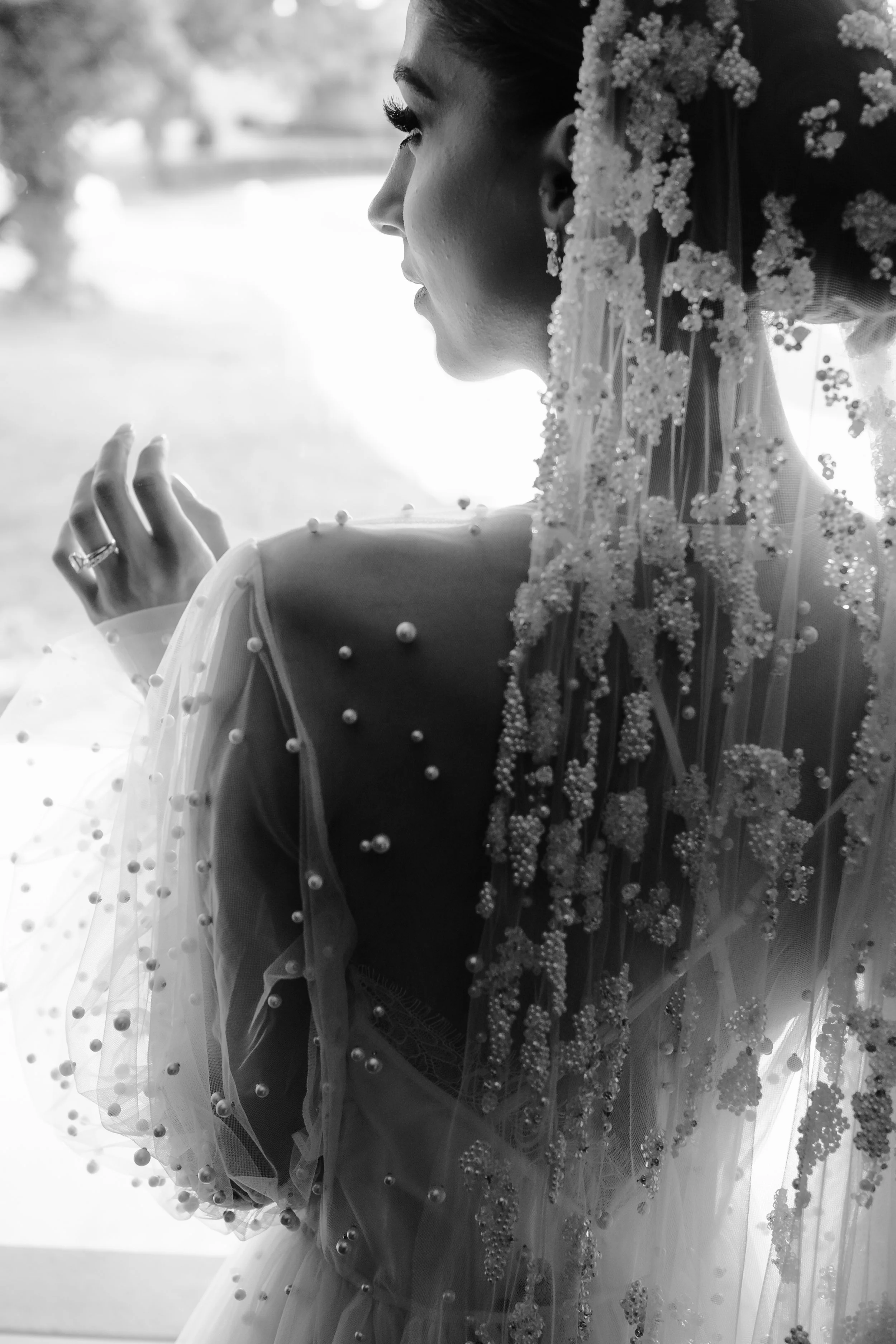 Black and white photo of a woman in a wedding dress and veil, standing near a window, looking to the side.