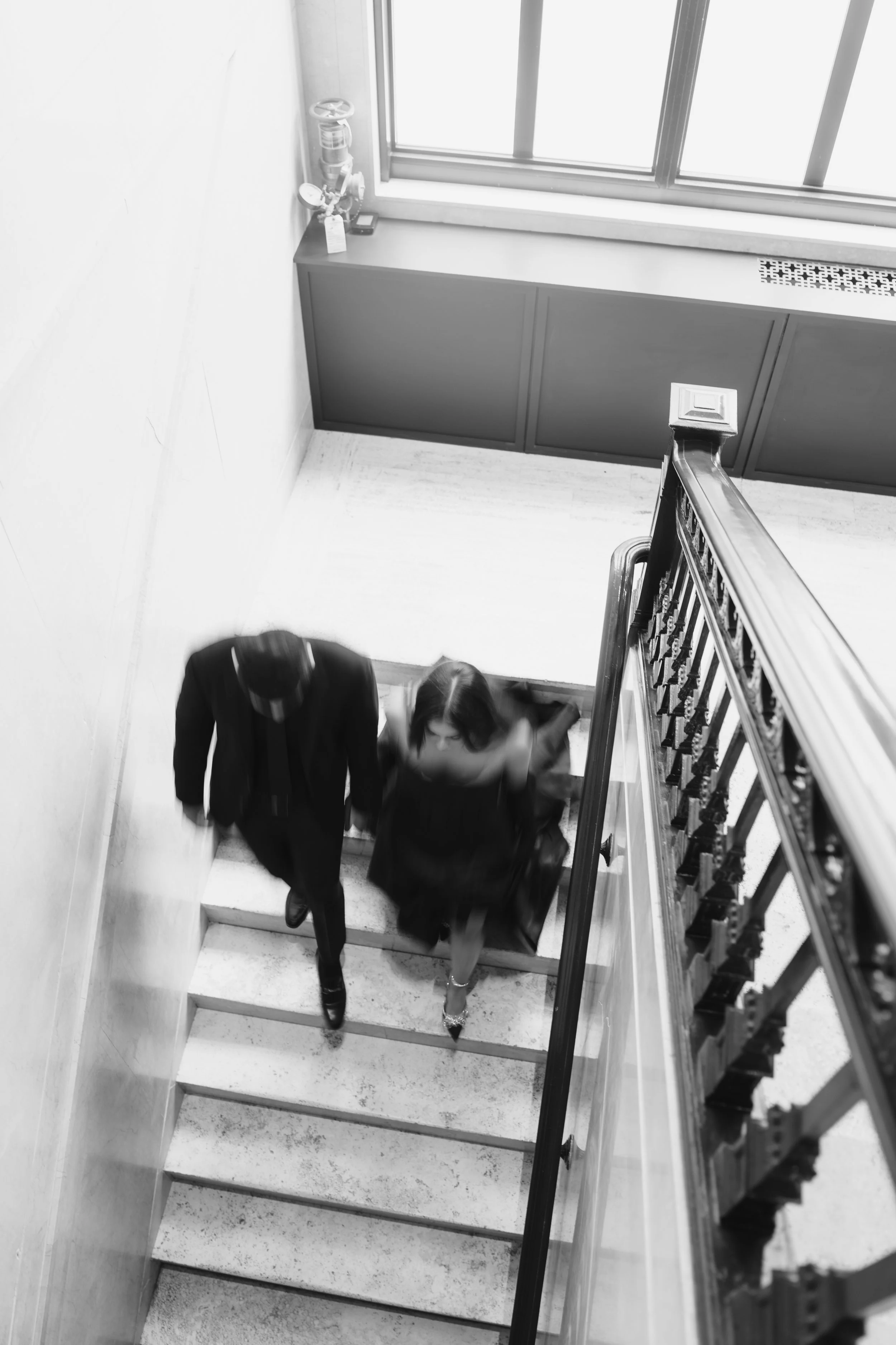 Black and white photo of a man and a woman walking up a staircase viewed from above. The man is dressed in a suit and the woman is wearing a dress and heels. The staircase has a decorative railing on the right and a window at the top of the staircase
