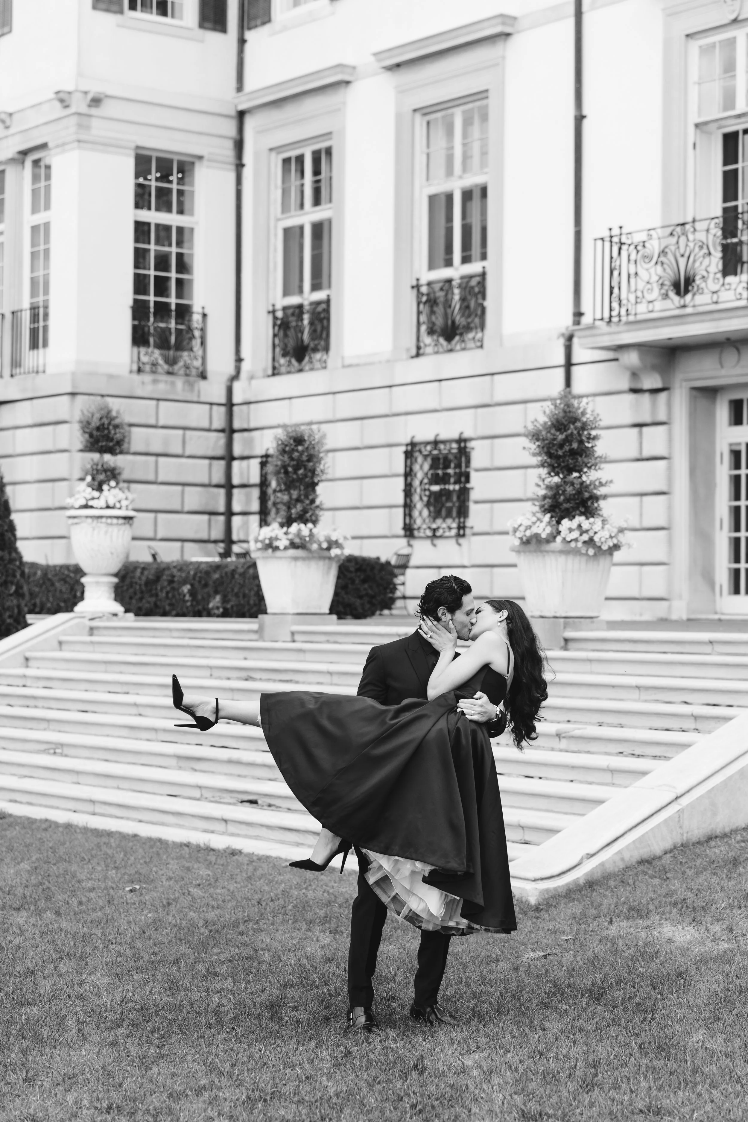 A man in a suit carrying a woman in a long dress and heels in front of a grand building with steps and large flower pots, black and white photograph.