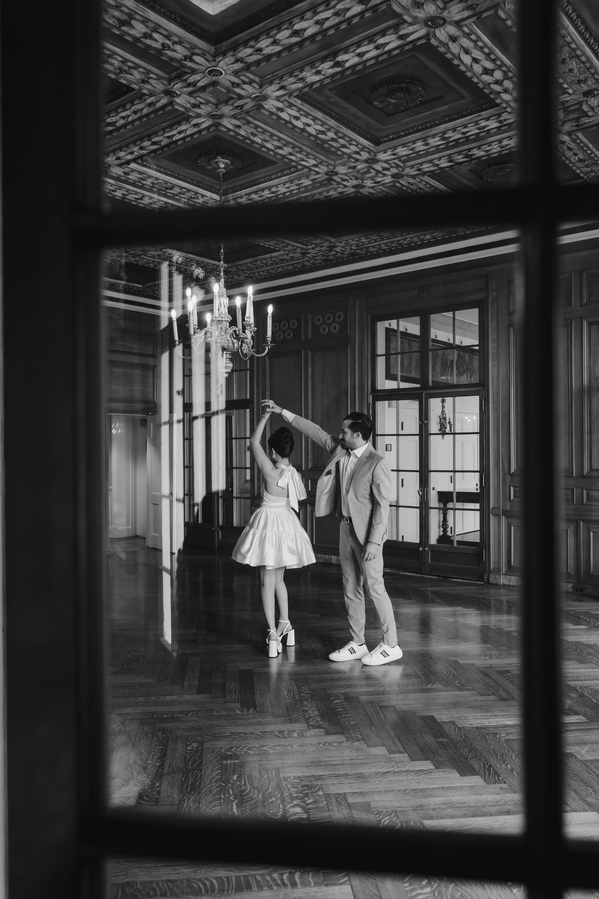 A man and woman dancing in an ornate, vintage-style room with wooden paneling, a chandelier, and large windows, viewed through a window frame in black and white.