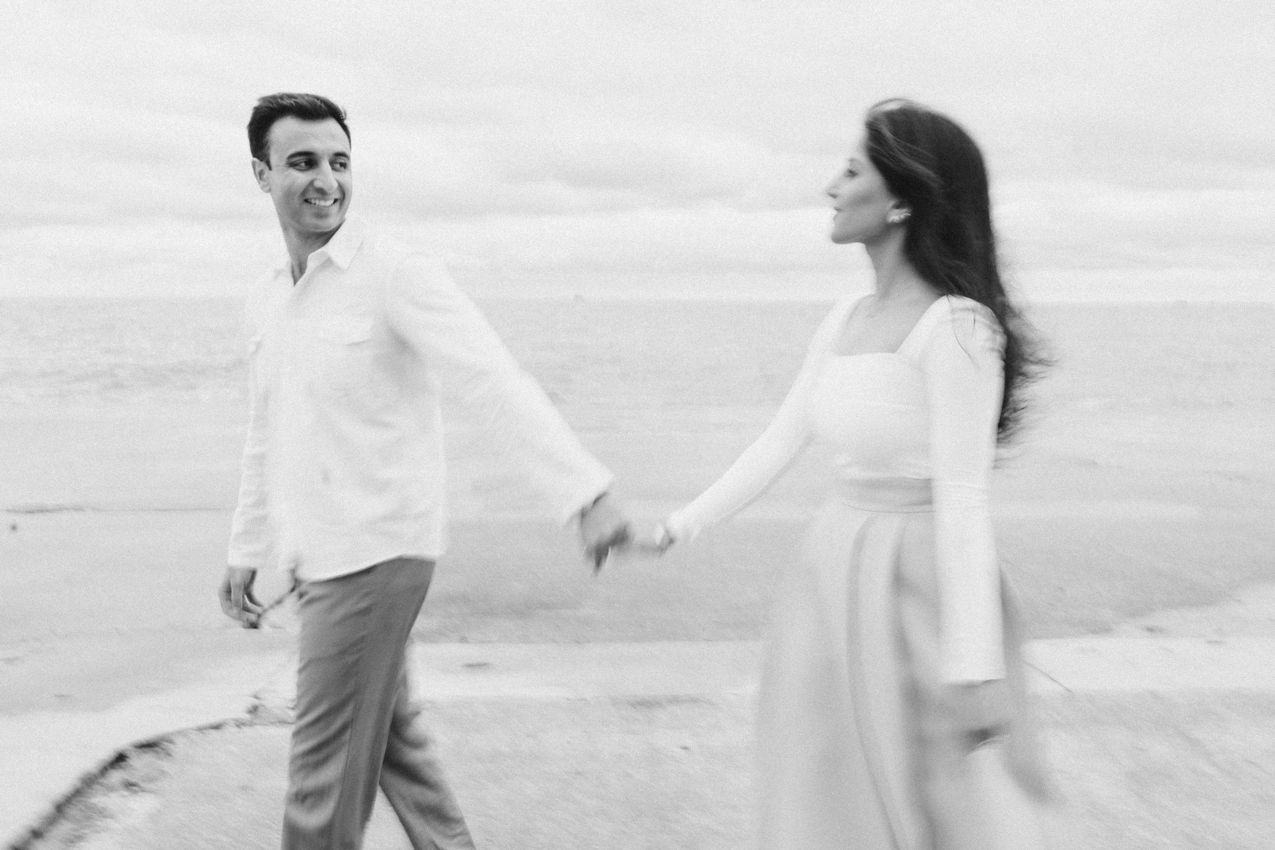 A black and white photo of a couple holding hands and walking along the beach, smiling and looking at each other.