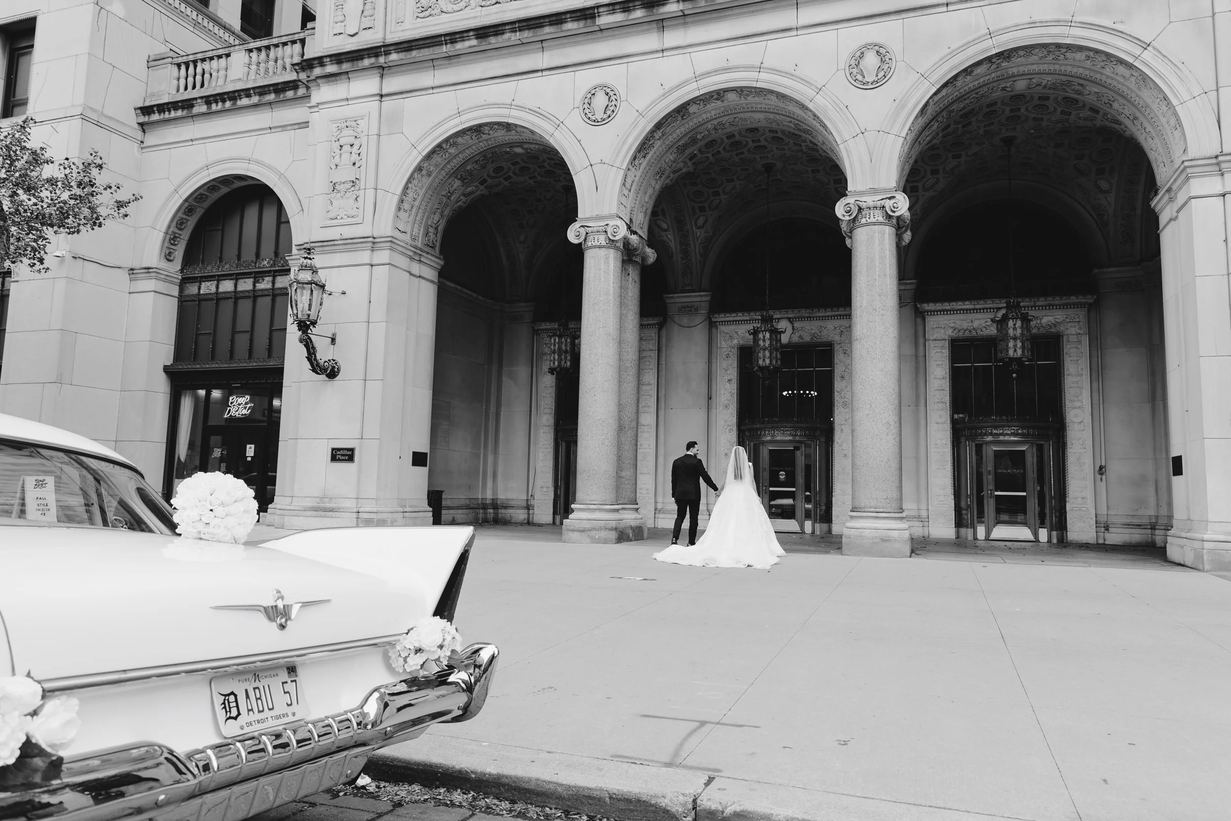 A bride and groom holding hands outside a large, classical-style building with arched entrances and columns. A vintage white car with floral decorations is parked in front.