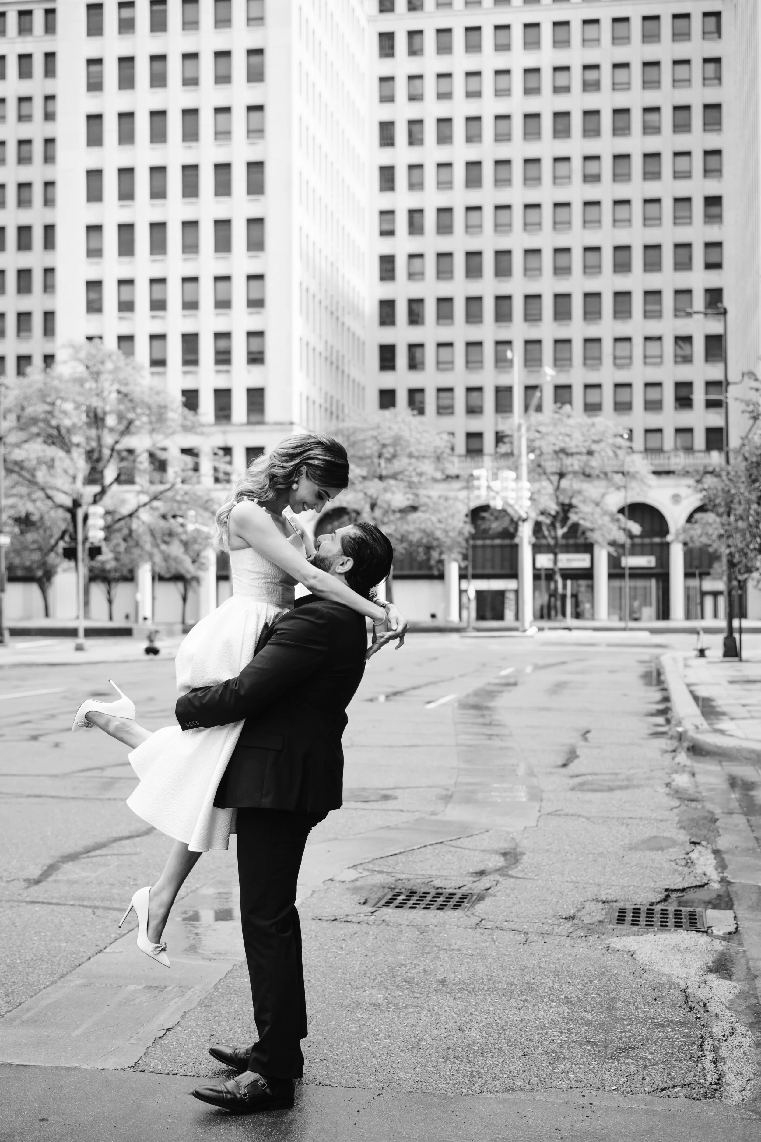 A man in a suit lifts a woman in a dress and high heels up in his arms on an empty city street with tall buildings and trees in the background.