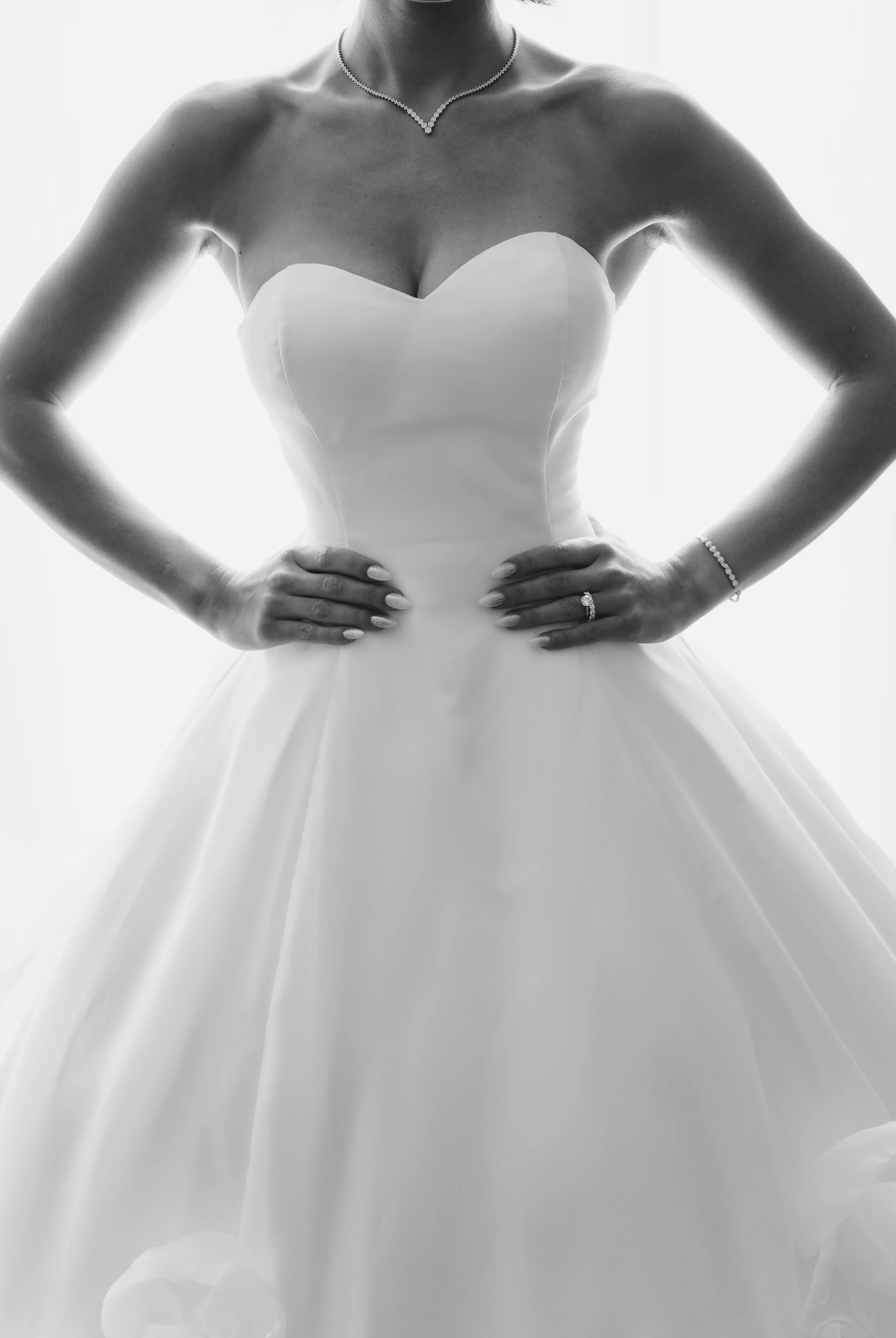 A woman in a strapless wedding dress, with her hands on her hips, wearing jewelry including a necklace, bracelet, and ring. The image is in black and white.