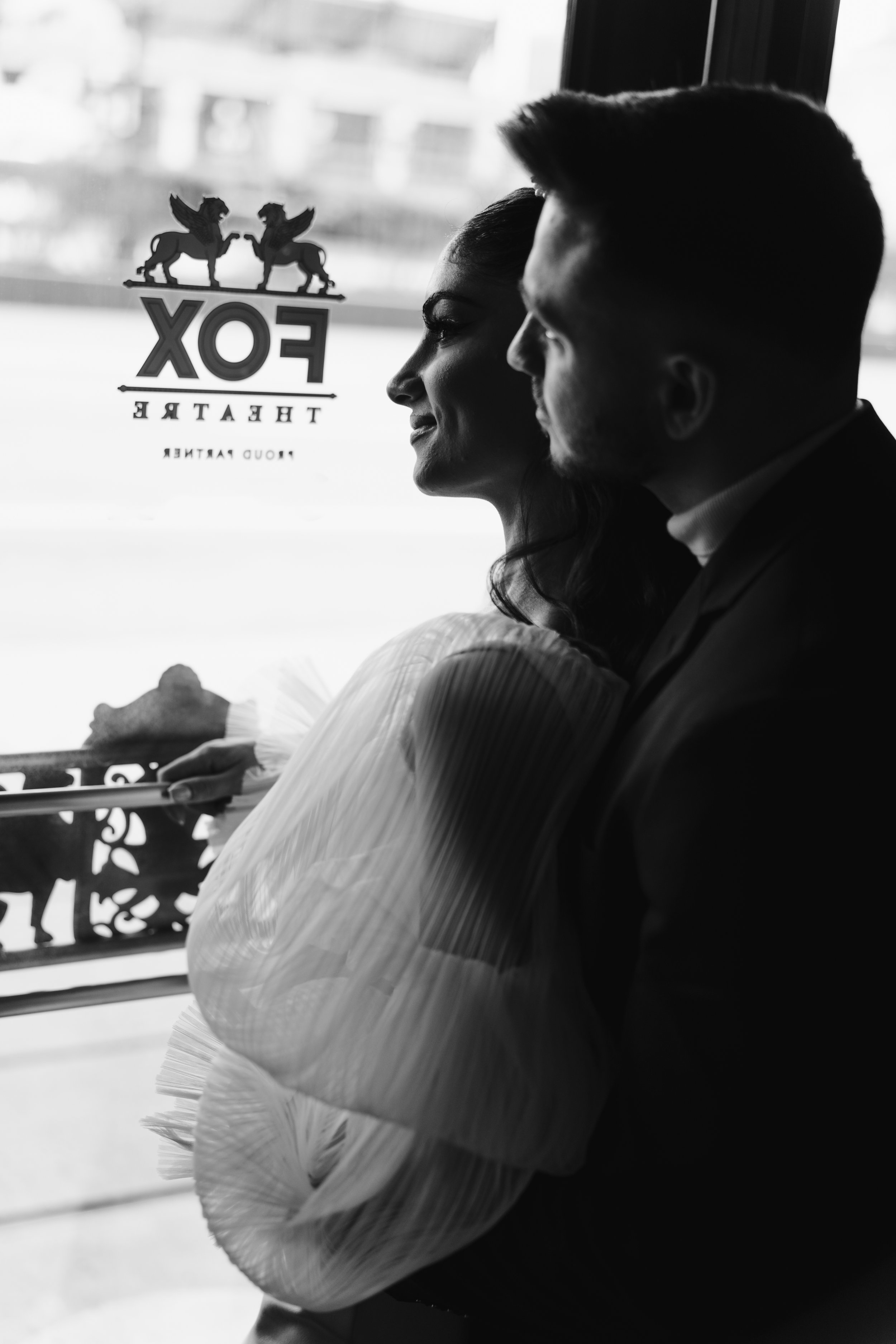 Black and white photo of a woman and a man sitting closely by a window, with the woman's arm crossed and holding a plate, looking outside. The window has a logo with lions and the words "FOX THEATRE" and "FROUD GARTNER" visible.