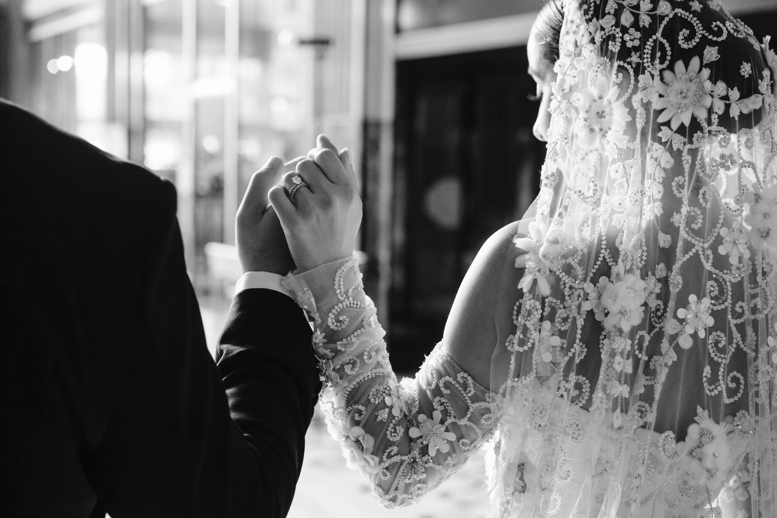 Black and white photo of a bride and groom holding hands during their wedding, with the bride wearing a lace dress with floral embroidery.