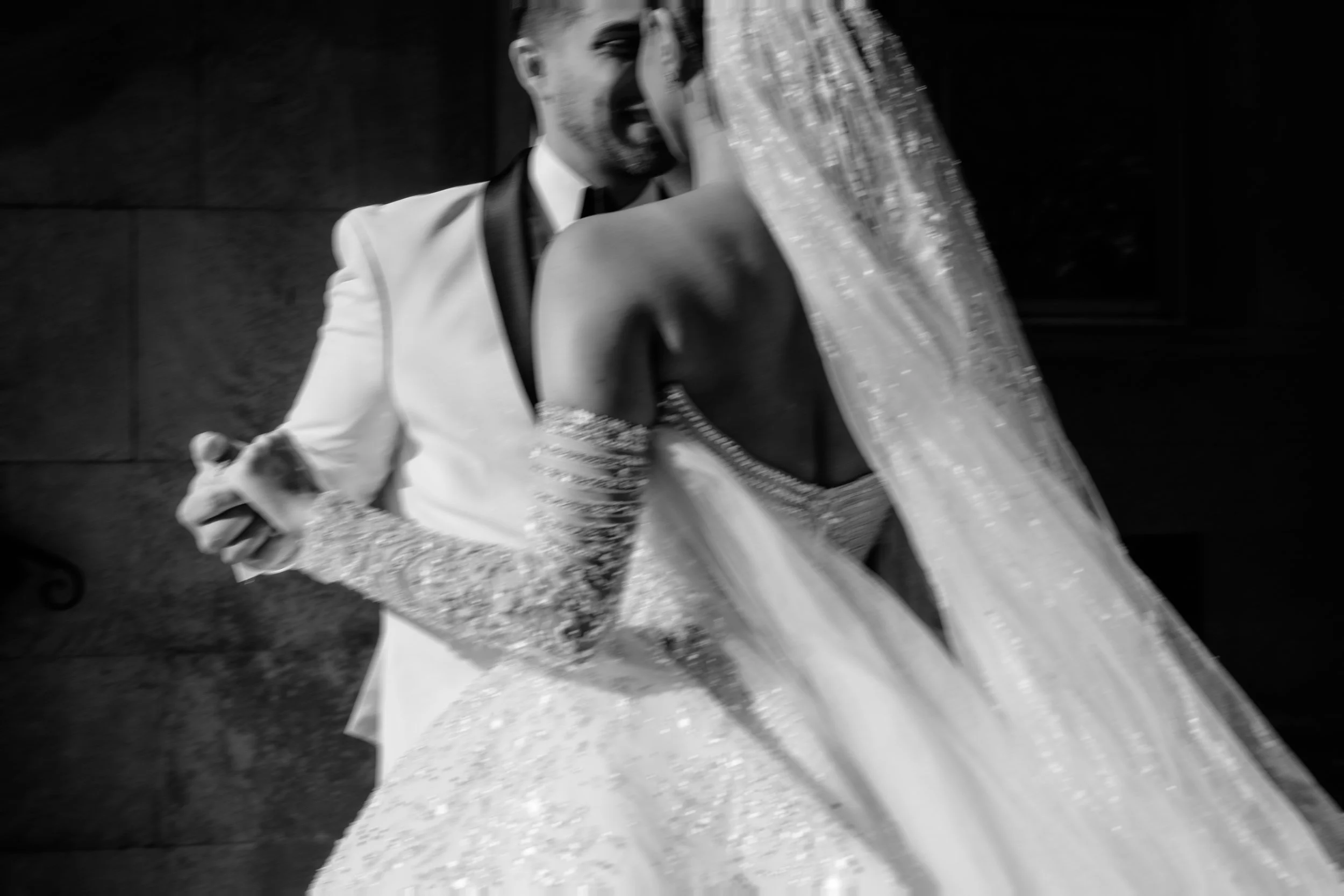 A black and white photo of a bride and groom dancing closely, with the groom wearing a tuxedo and the bride in a sparkling gown with a veil.