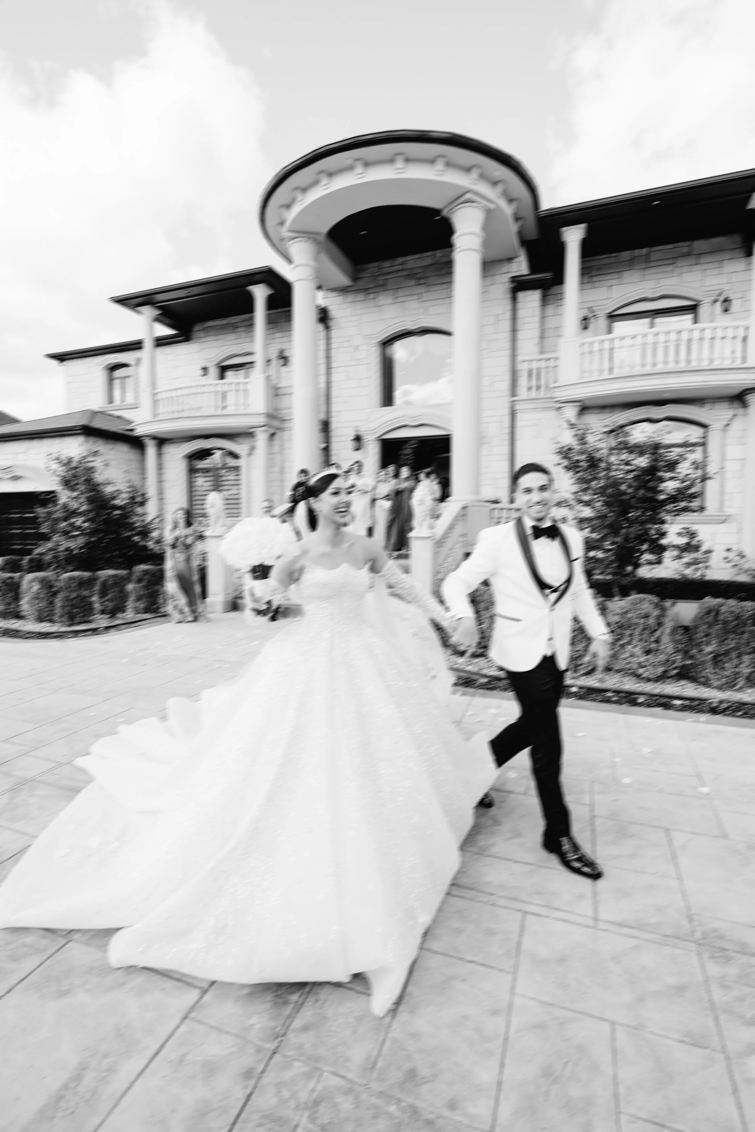Black and white photo of a bride and groom running out of a large house, with guests in the background, on a sunny day.