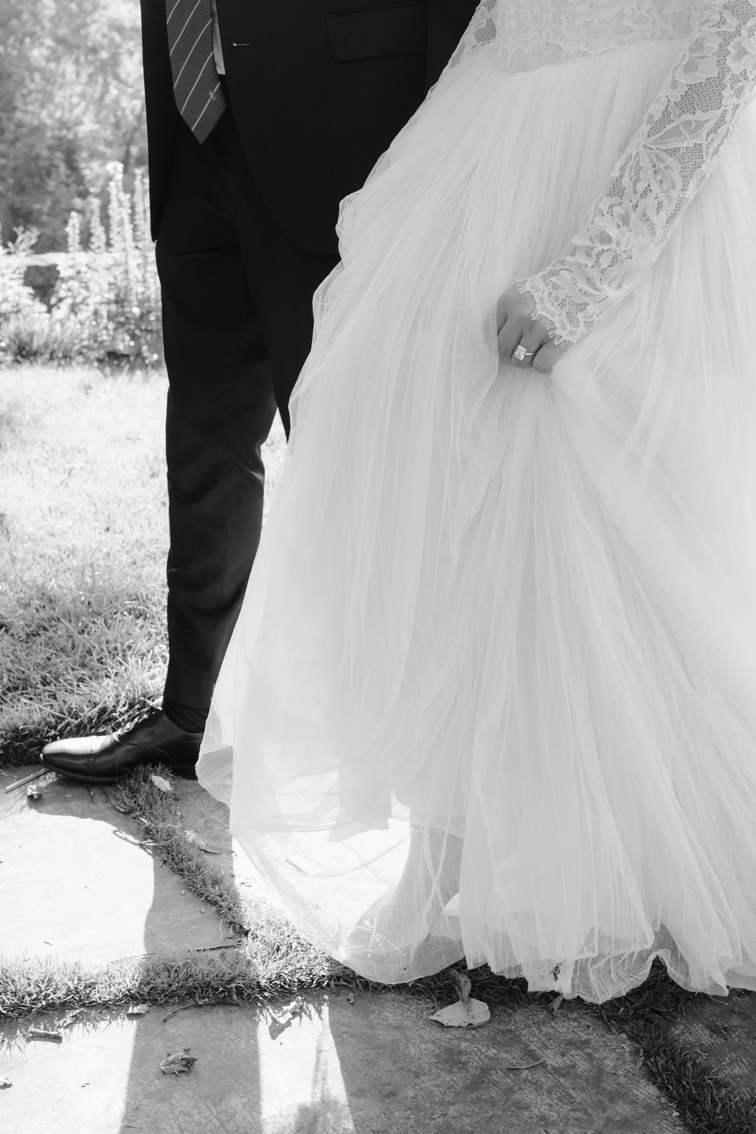 Close-up of a bride in a white wedding dress and a groom in a black suit standing outdoors, with focus on the bride's hand showing a wedding ring.