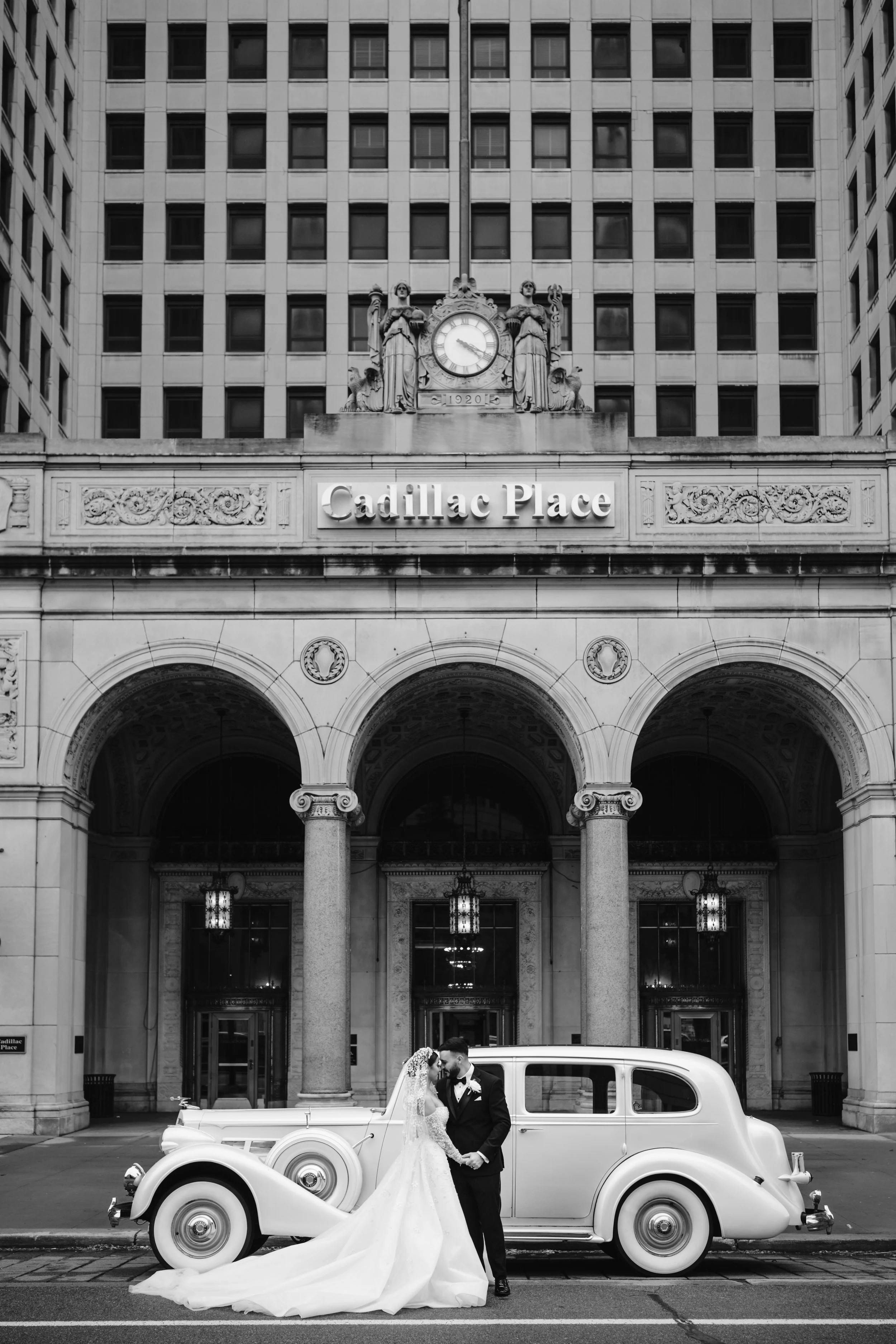 A newlywed couple in wedding attire standing in front of a vintage car on a city street, with a building labeled 'Cadillac Place' in the background.