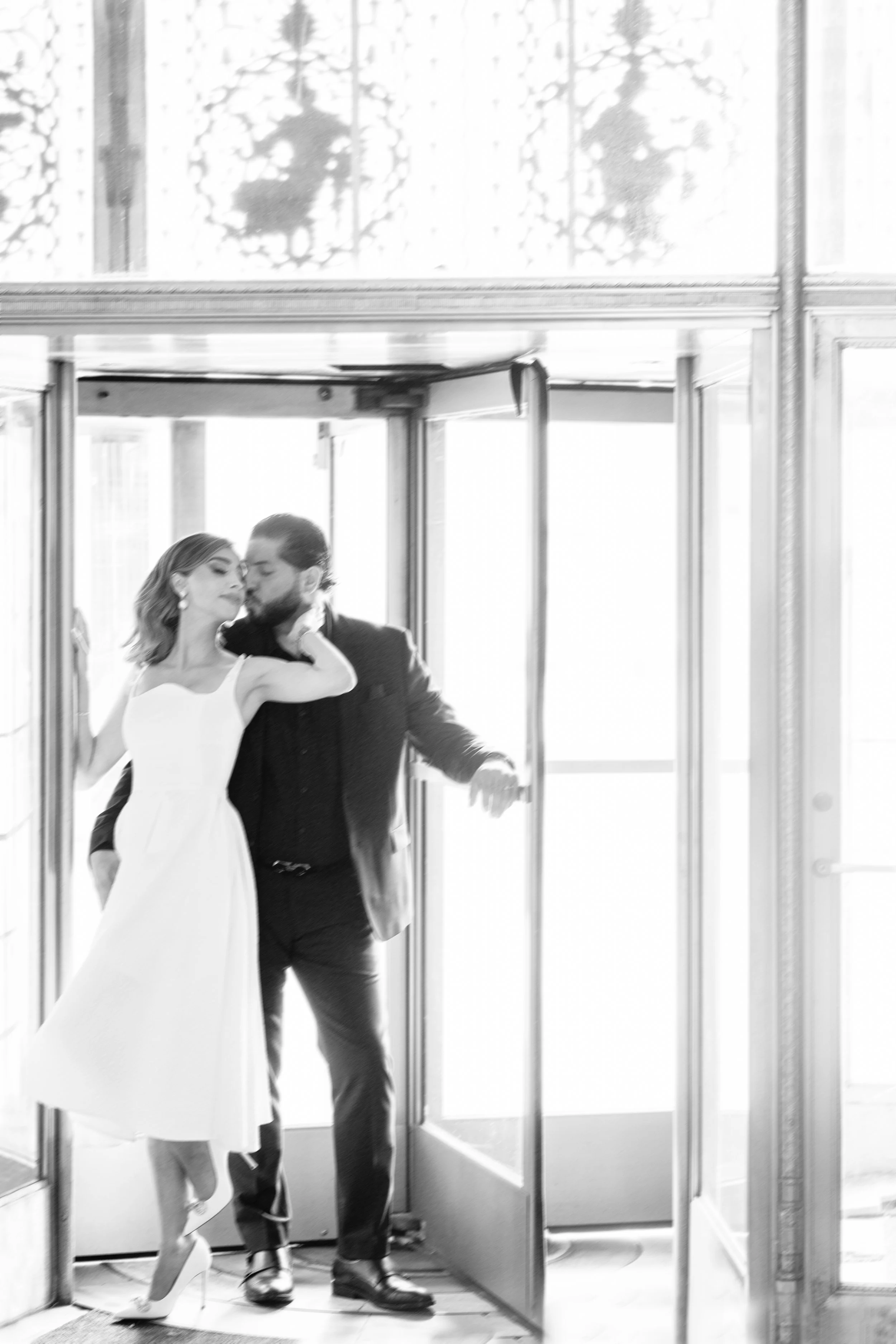 A couple sharing a kiss in an open-door entrance, with the woman wearing a white dress and high heels, and the man in a dark suit, in black and white photography.