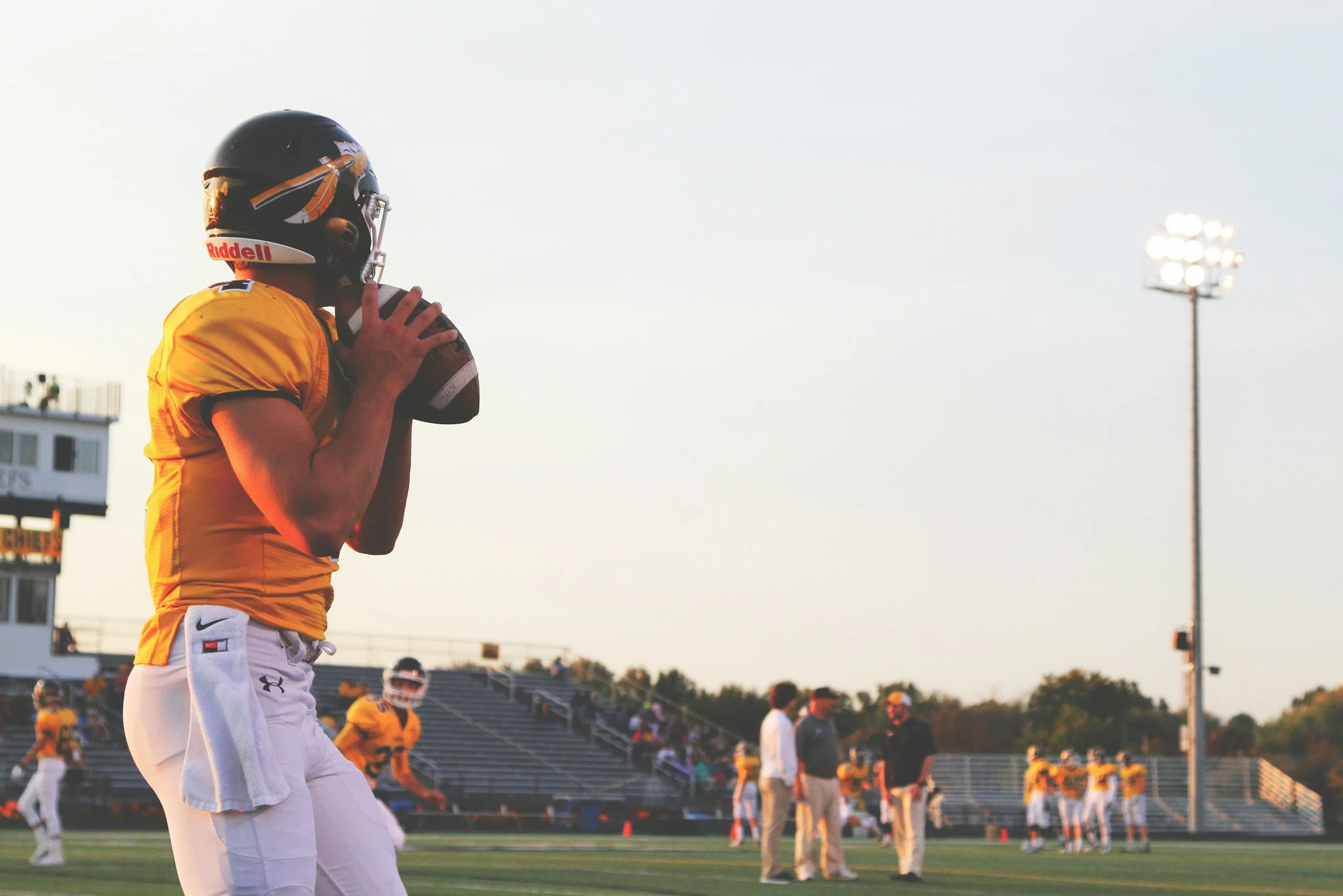 A football player wearing a yellow jersey and white pants is preparing to throw a football on a field during sunset, with team members, coaches, and spectators in the background.