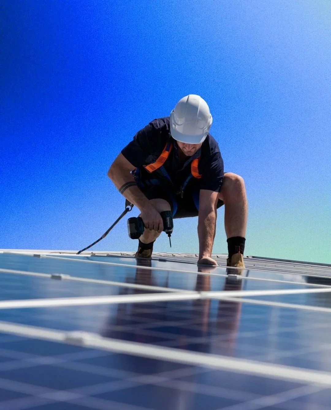 Worker installing solar panels on a roof, wearing a white safety helmet, black shorts, and a black shirt, with a blue sky in the background.