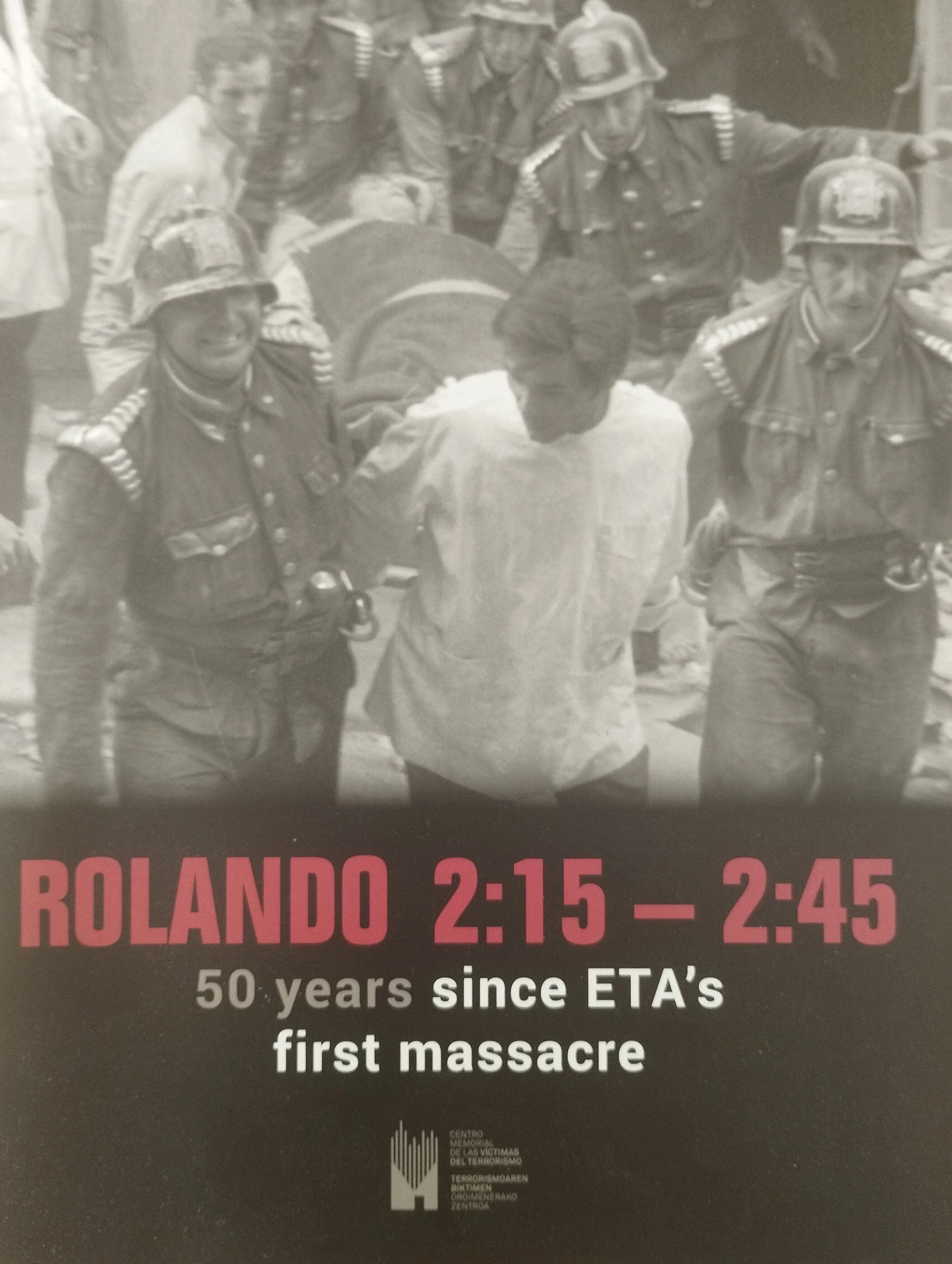 Cartel conmemorativo en tonos sepia y rosa, con una foto en blanco y negro de policías y una víctima. Texto que dice: "Rolando 2:15 - 2:45, 50 años desde la primera masacre de ETA."