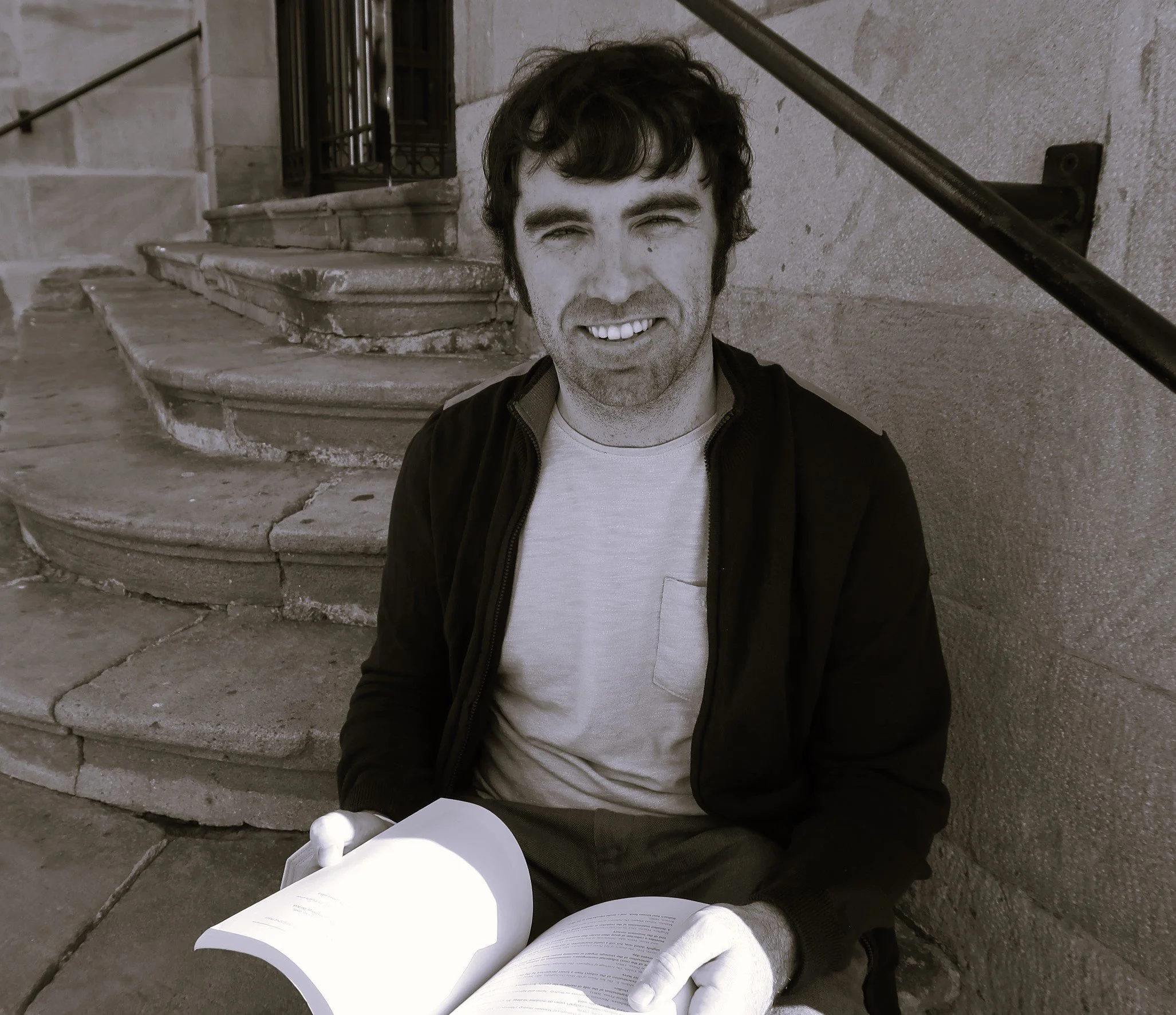 Niall Cullen, PhD - Academic English Servicess. Traductor académico. Sonriendo, sentado en las escaleras de piedra leyendo un libro, en un entorno urbano.