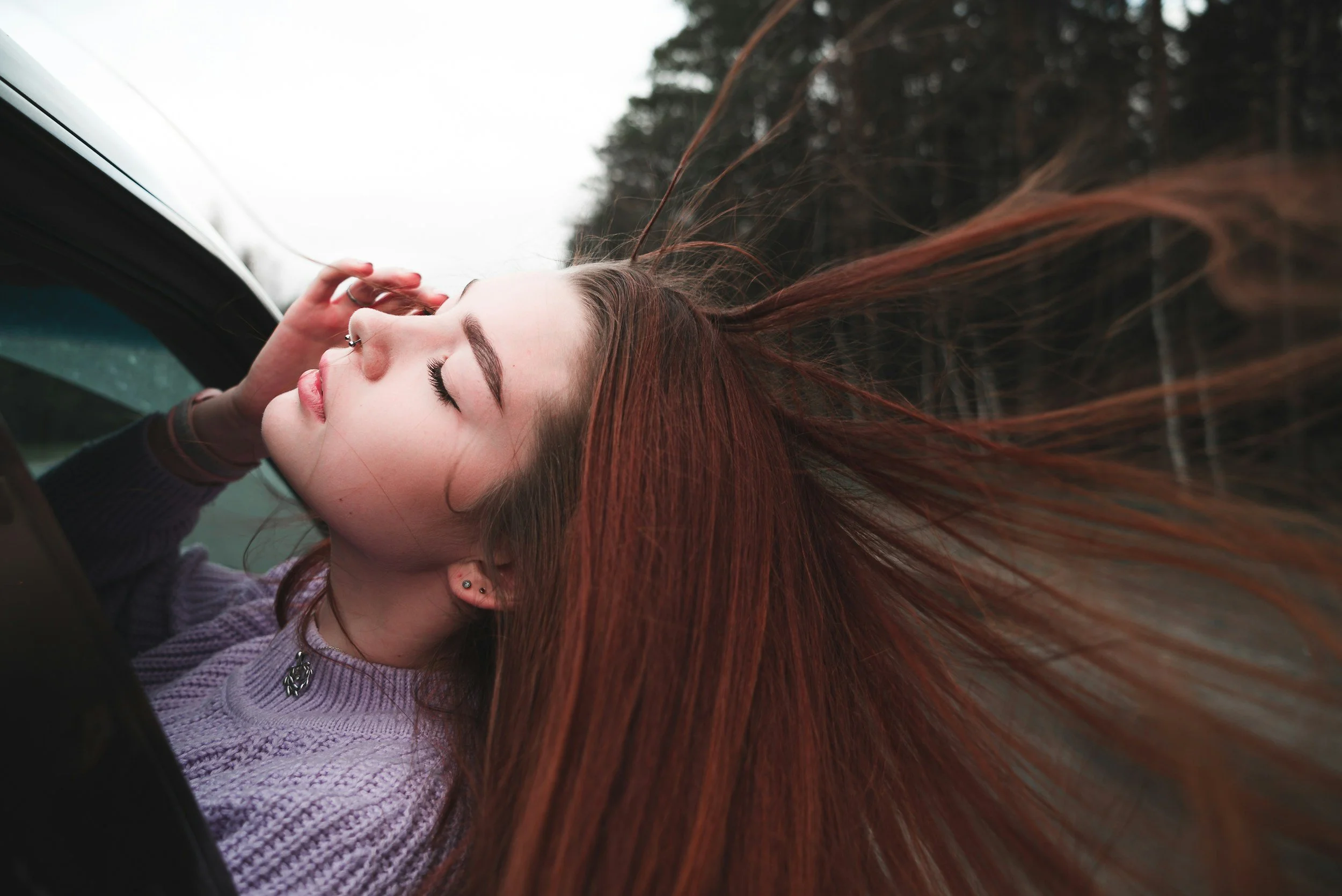 A young woman with red hair leaning out of a car window with her eyes closed, hair blowing in the wind, in a forested area.