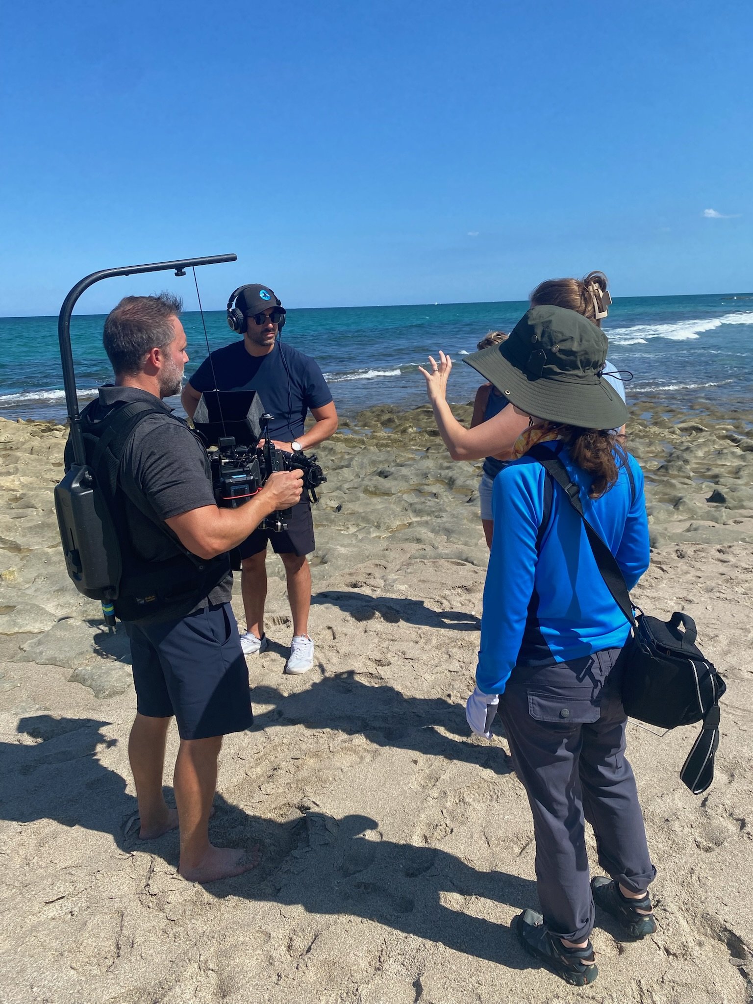 A group of five people standing on a sandy beach, with the ocean and a clear blue sky in the background. One person is holding a camera, others are talking or listening, two wearing headphones and hats.