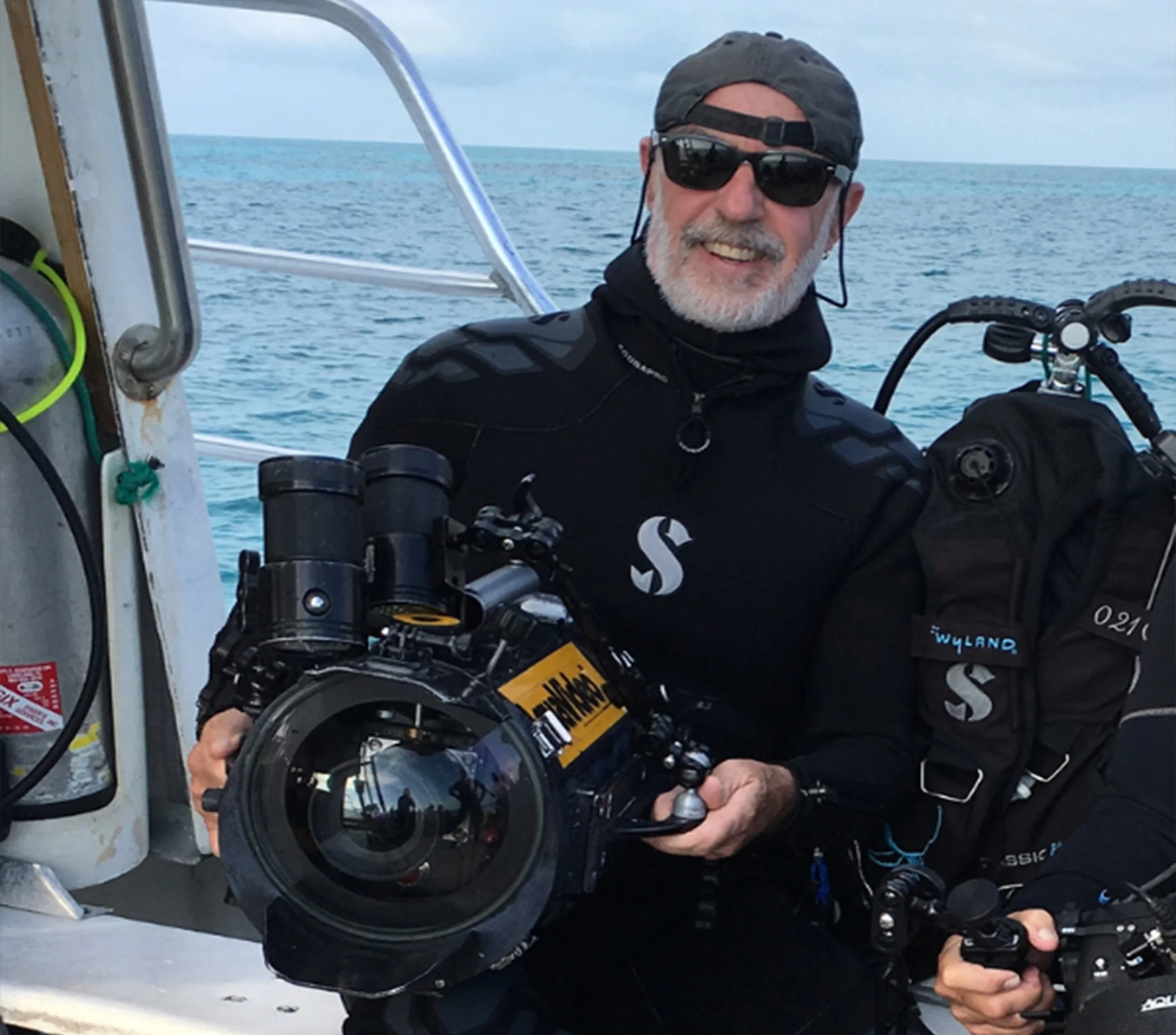 A man with gray beard wearing sunglasses, a cap backwards, and black clothing, holding a camera or device on a boat with a view of the ocean in the background.