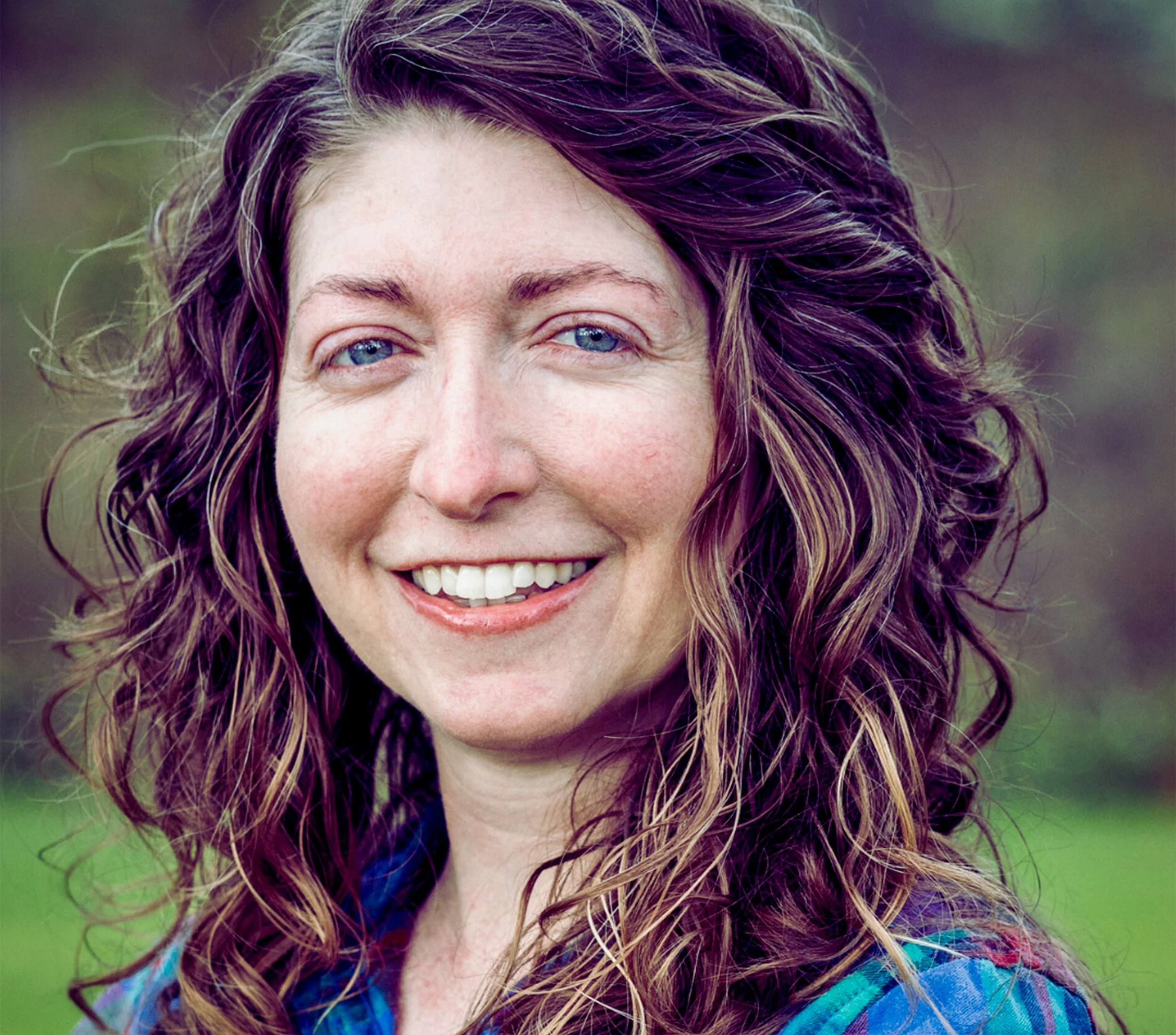 Close-up of a woman with curly hair, blue eyes, smiling, outdoors with blurred green background.